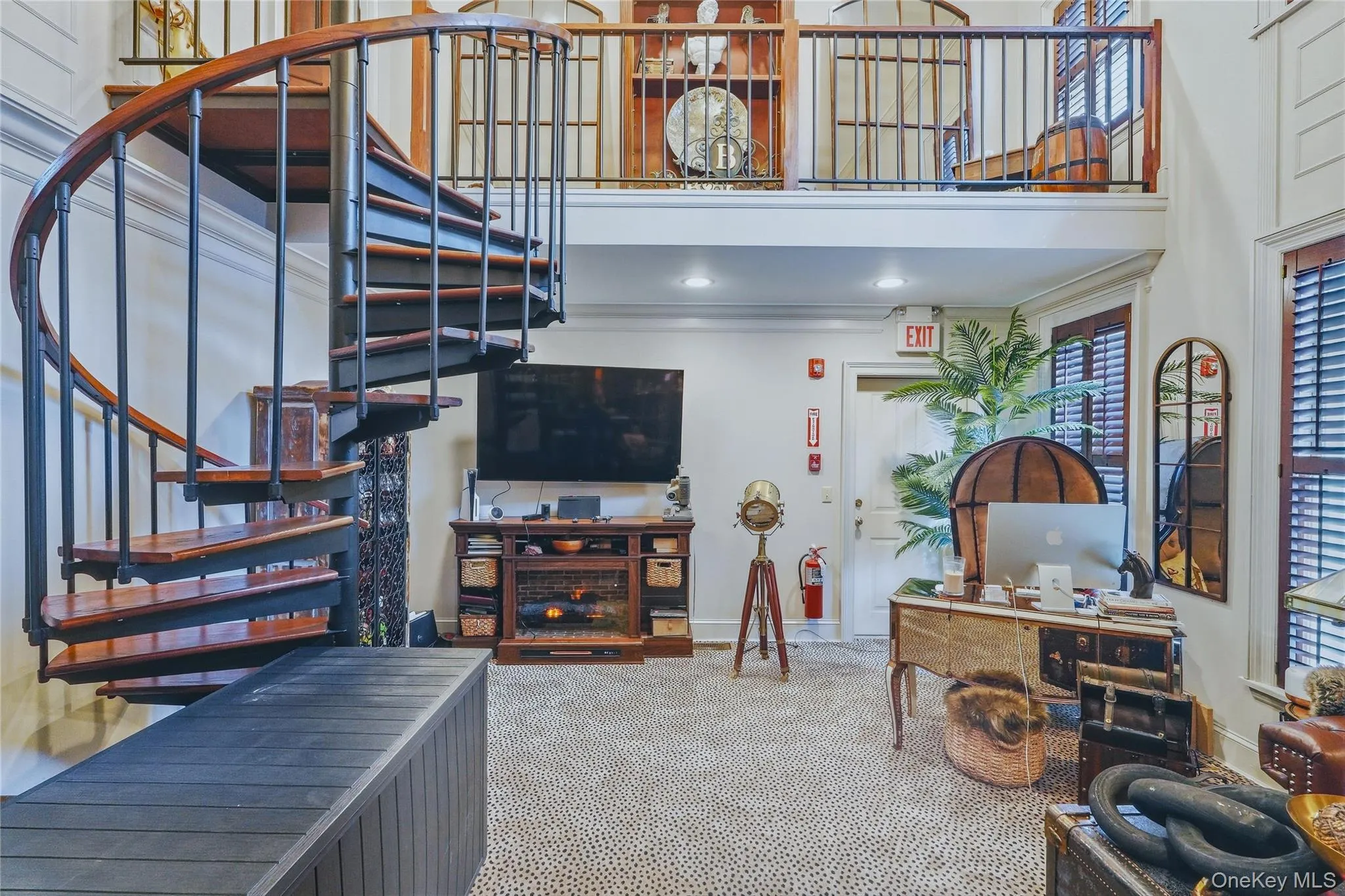 Spiral staircase with wood treads and metal balusters, upper-level balcony with metal railing, built-in media console, interior shutters on windows, and a carpeted floor Spiral staircase with wood treads and metal balusters, upper-level balcony with metal railing, built-in media console, interior shutters on windows, and a carpeted floor