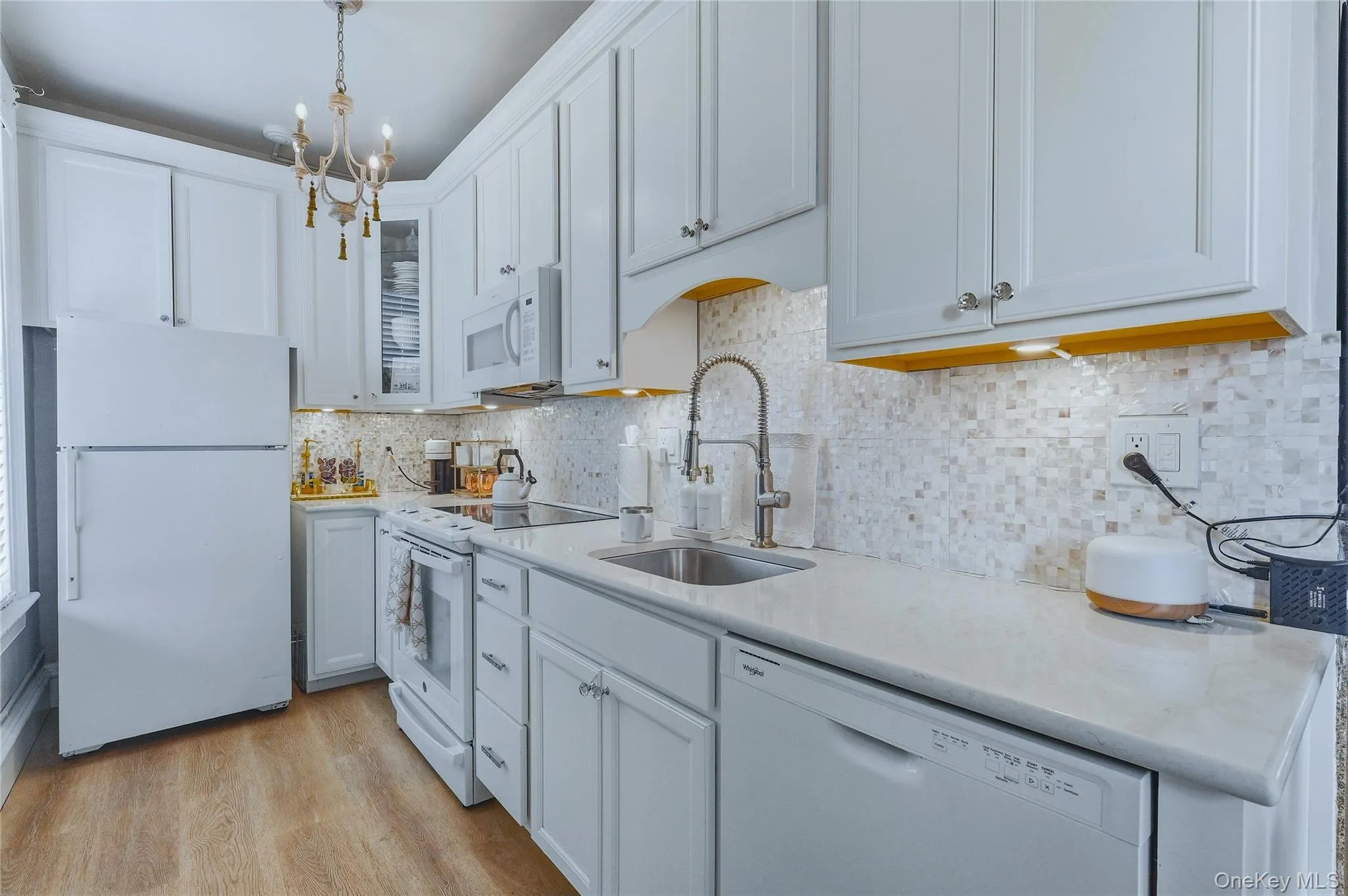White kitchen cabinetry, stainless steel sink with a high-arc faucet, mosaic tile backsplash, light-colored countertops, and wood-finish flooring White kitchen cabinetry, stainless steel sink with a high-arc faucet, mosaic tile backsplash, light-colored countertops, and wood-finish flooring