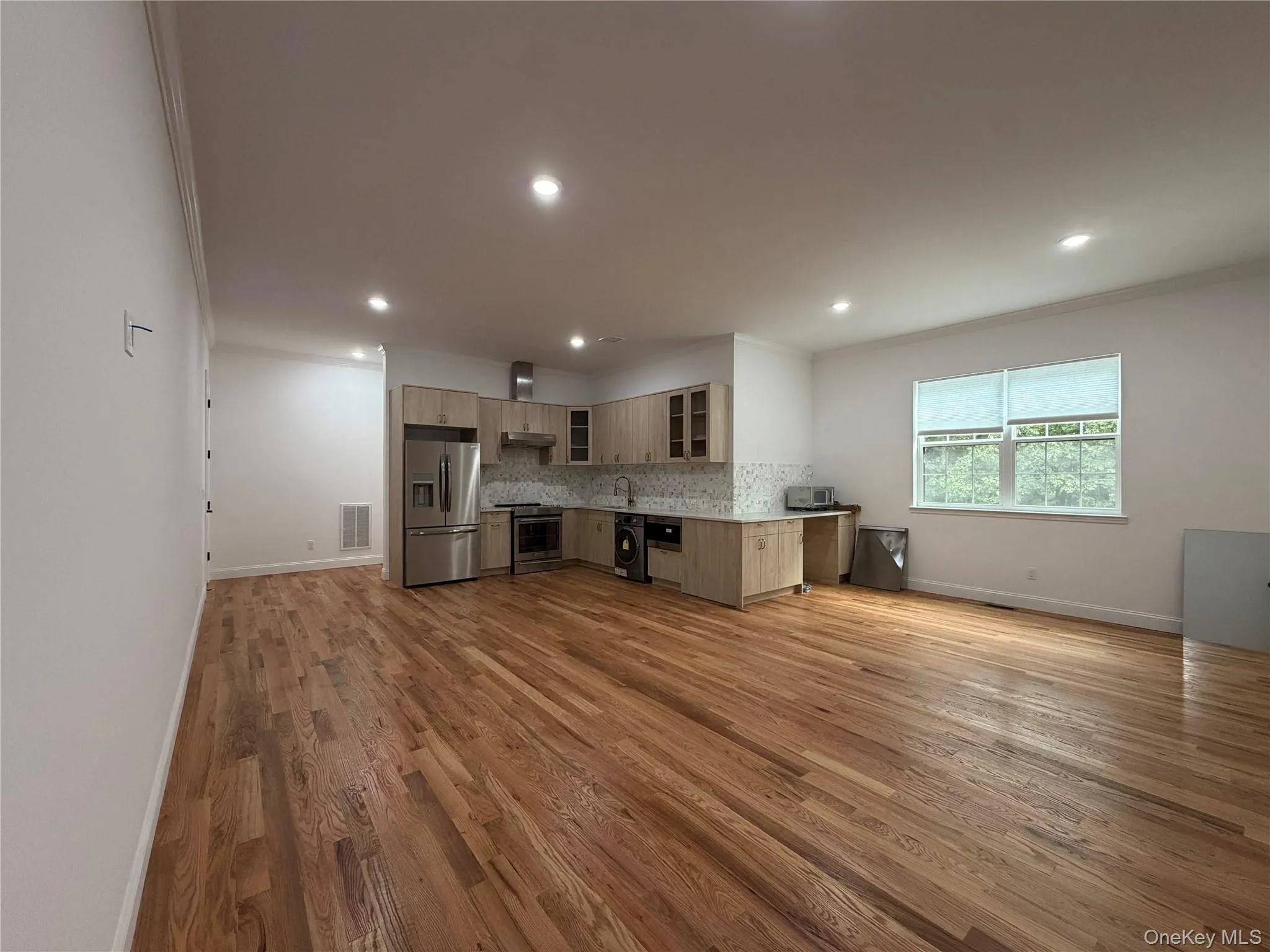 Open-concept living area featuring hardwood flooring, recessed lighting, and a kitchen with stainless steel appliances, light wood-finish cabinetry, and a full tile backsplash Open-concept living area featuring hardwood flooring, recessed lighting, and a kitchen with stainless steel appliances, light wood-finish cabinetry, and a full tile backsplash