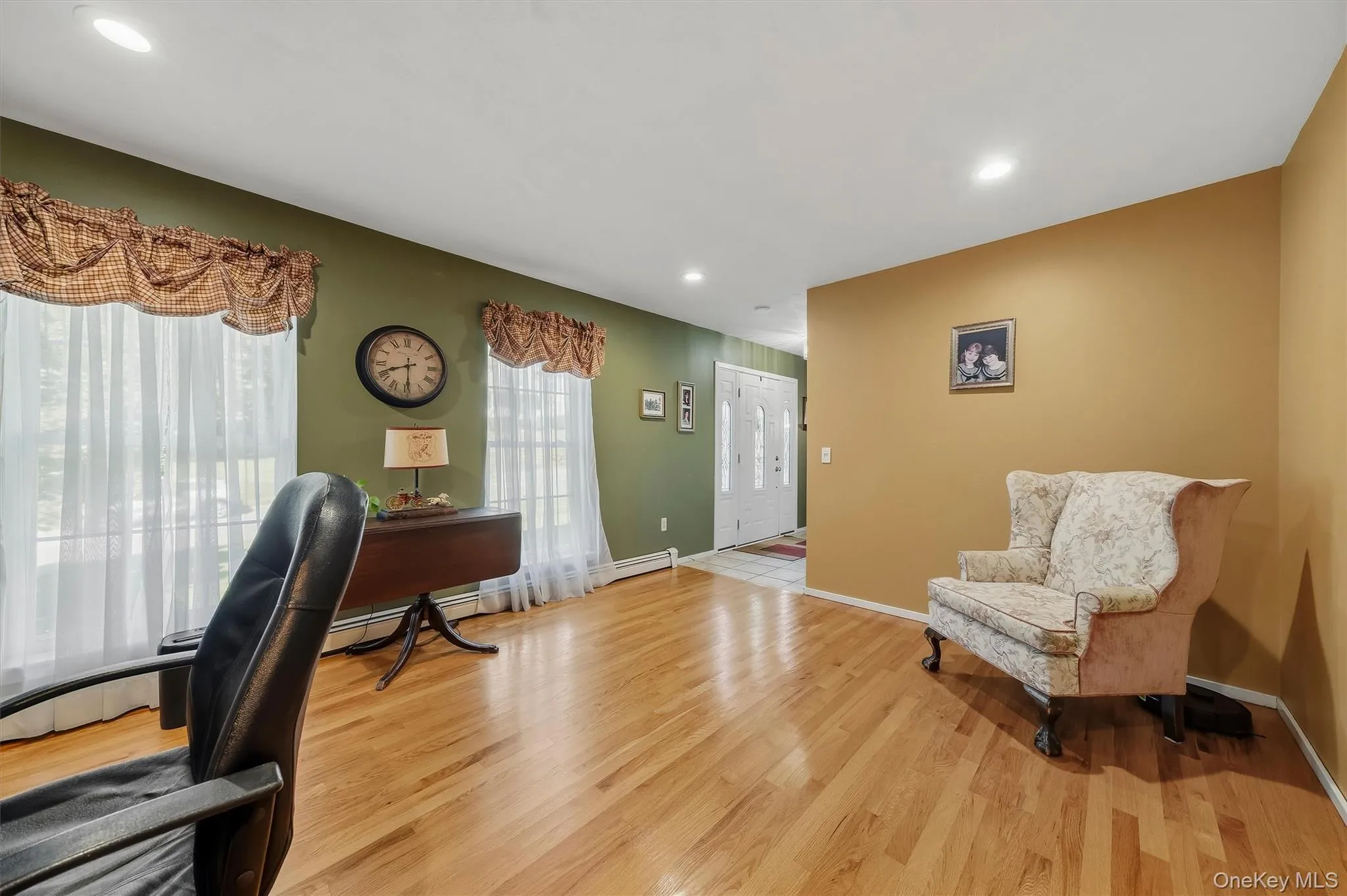 Spacious home office featuring warm wood-finish flooring, dual windows with decorative valances and sheer panels, green accent wall with wall-mounted clock, and a complementary upholstered armchair Spacious home office featuring warm wood-finish flooring, dual windows with decorative valances and sheer panels, green accent wall with wall-mounted clock, and a complementary upholstered armchair