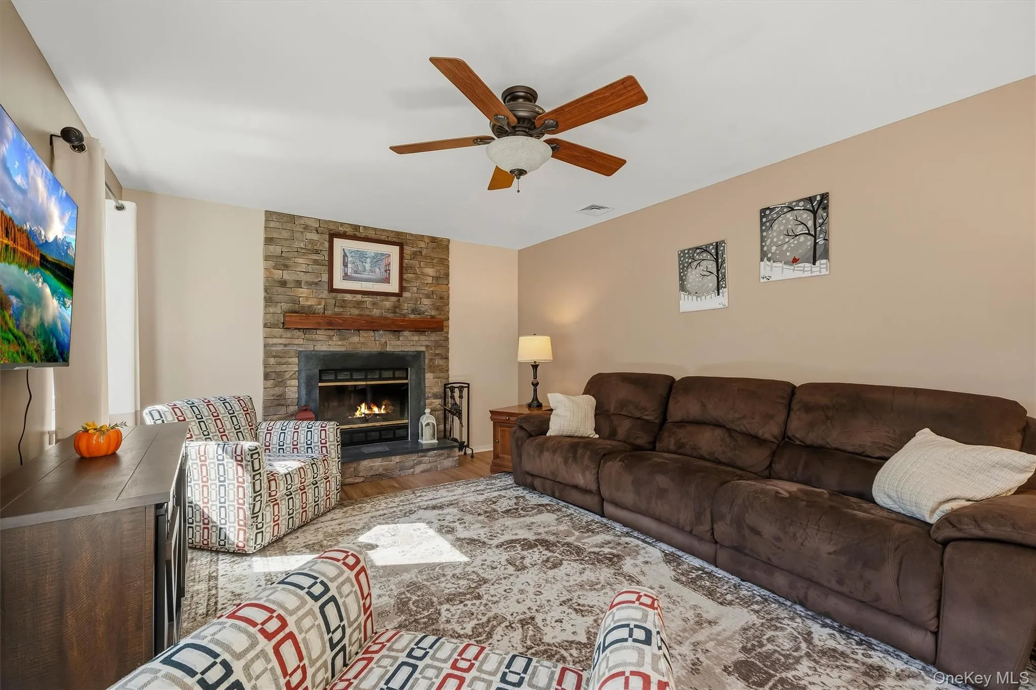 Stone-surround fireplace with a wood mantle, ceiling fan with integrated lighting, recessed lighting, and warm neutral wall tones Stone-surround fireplace with a wood mantle, ceiling fan with integrated lighting, recessed lighting, and warm neutral wall tones