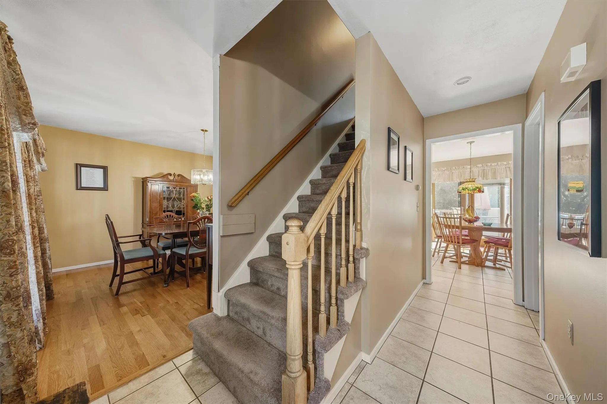 Hallway with square tile flooring, staircase with carpeted treads and wood banister, and wood-finish flooring in an adjoining room Hallway with square tile flooring, staircase with carpeted treads and wood banister, and wood-finish flooring in an adjoining room