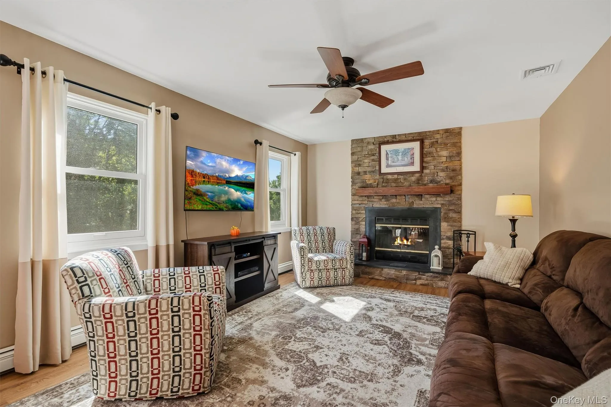Stone-faced fireplace with a wood mantel, wood-finish flooring, and a ceiling fan Stone-faced fireplace with a wood mantel, wood-finish flooring, and a ceiling fan
