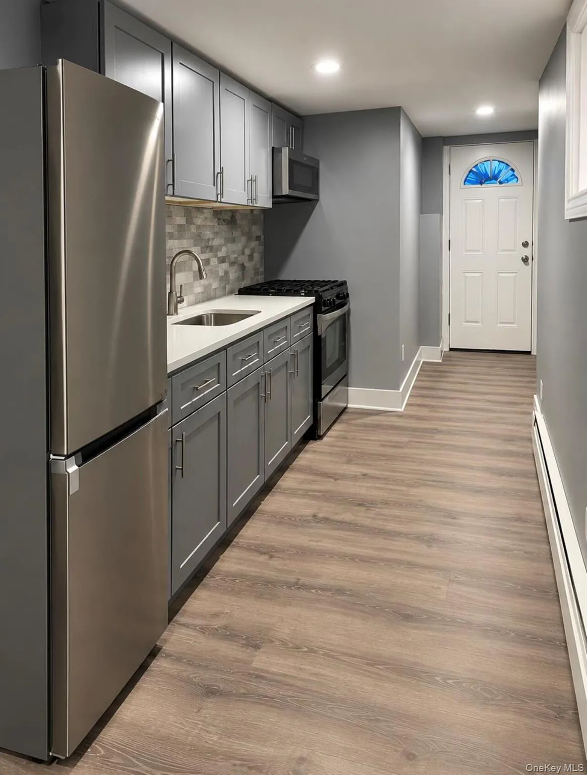 Galley kitchen featuring stainless steel appliances, grey cabinetry, mosaic tile backsplash, and wood-finish flooring Galley kitchen featuring stainless steel appliances, grey cabinetry, mosaic tile backsplash, and wood-finish flooring