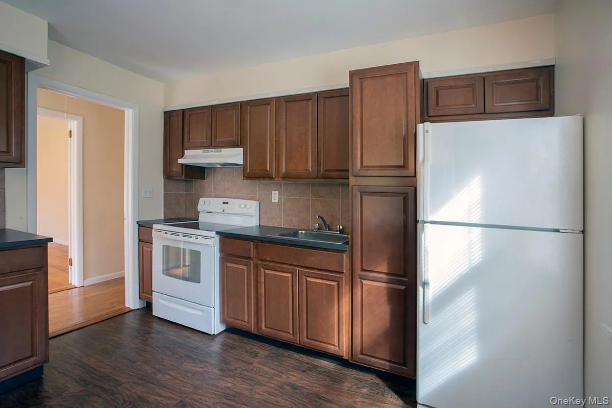 Kitchen featuring dark wood-finish cabinetry, white appliances, dark countertops, and tile backsplash Kitchen featuring dark wood-finish cabinetry, white appliances, dark countertops, and tile backsplash