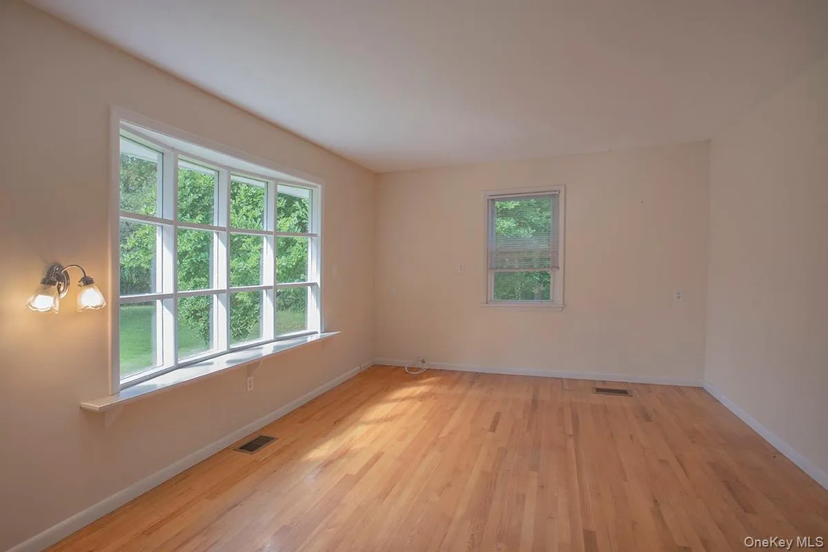 Hardwood flooring throughout, large bay window with white trim, single window with white trim, and wall-mounted sconce lighting Hardwood flooring throughout, large bay window with white trim, single window with white trim, and wall-mounted sconce lighting