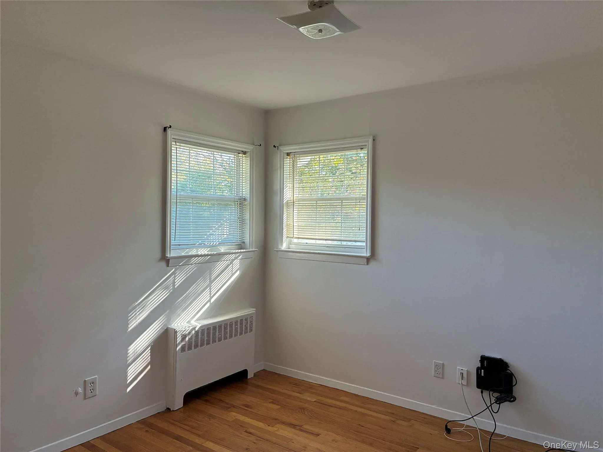 Hardwood flooring, dual windows with blinds, baseboard heating unit, and a ceiling-mounted light fixture Hardwood flooring, dual windows with blinds, baseboard heating unit, and a ceiling-mounted light fixture