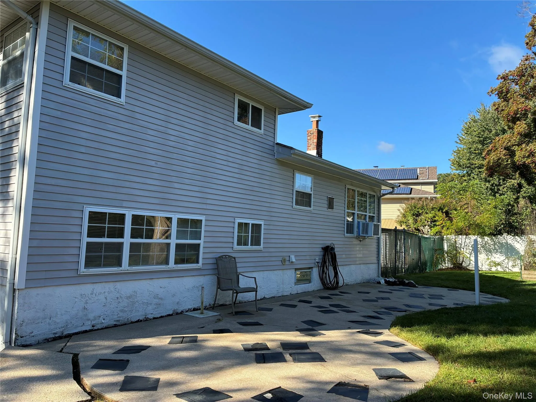 Rear exterior featuring light-colored siding, a brick chimney, multiple windows, a concrete patio with inset dark pavers, and a manicured lawn Rear exterior featuring light-colored siding, a brick chimney, multiple windows, a concrete patio with inset dark pavers, and a manicured lawn