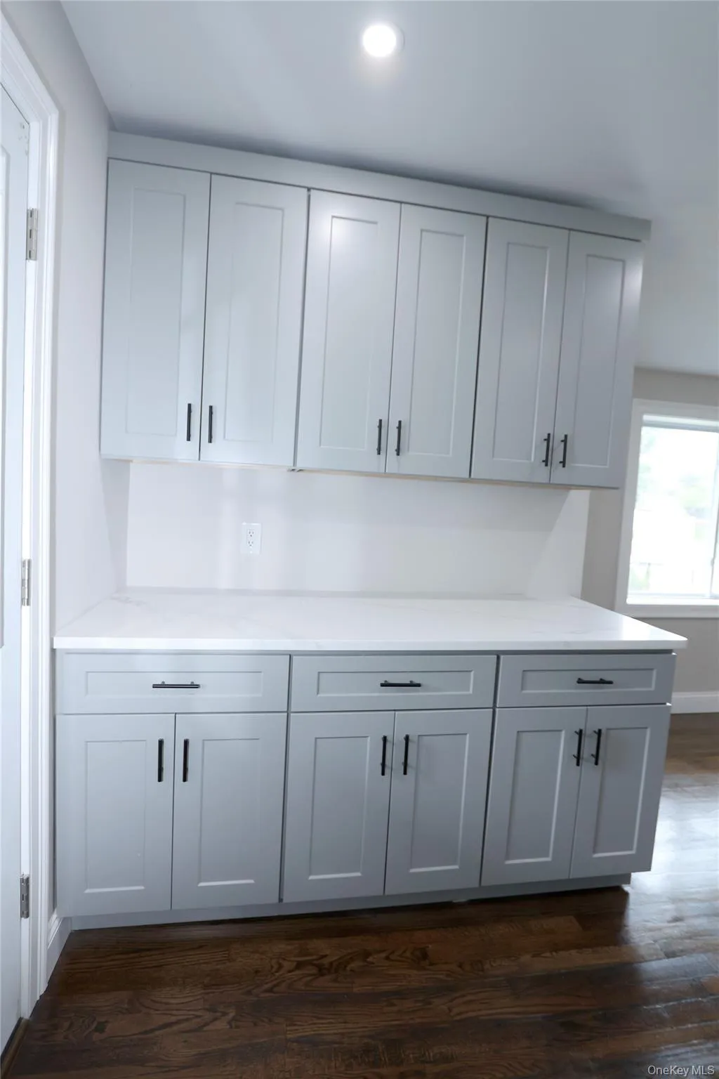 Light gray shaker-style cabinetry with black hardware, white countertop, and dark wood-finish flooring Light gray shaker-style cabinetry with black hardware, white countertop, and dark wood-finish flooring