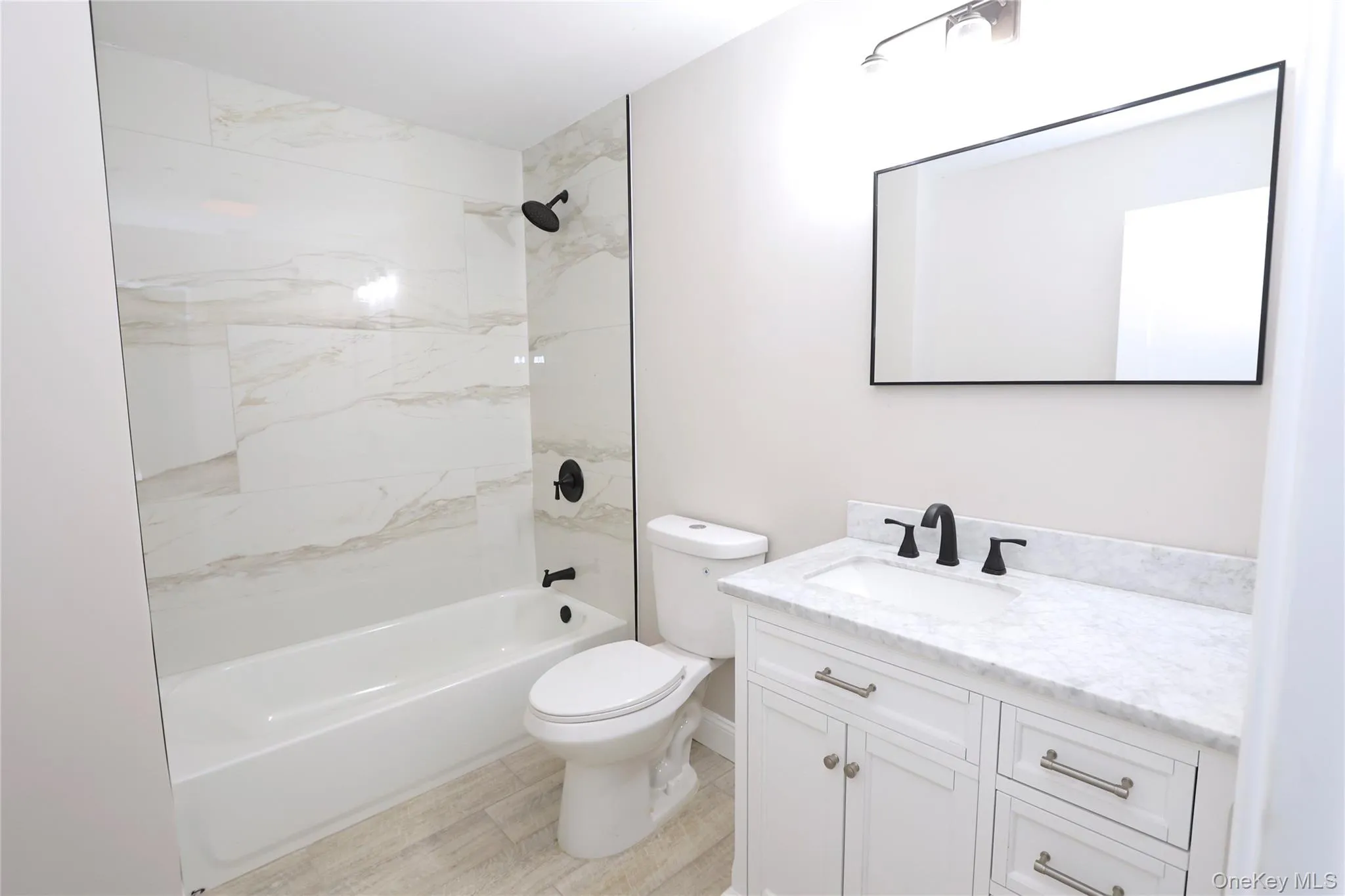 White tile bathtub with matte black fixtures, glass panel, wood-finish flooring, white vanity with stone countertop, and rectangular mirror White tile bathtub with matte black fixtures, glass panel, wood-finish flooring, white vanity with stone countertop, and rectangular mirror