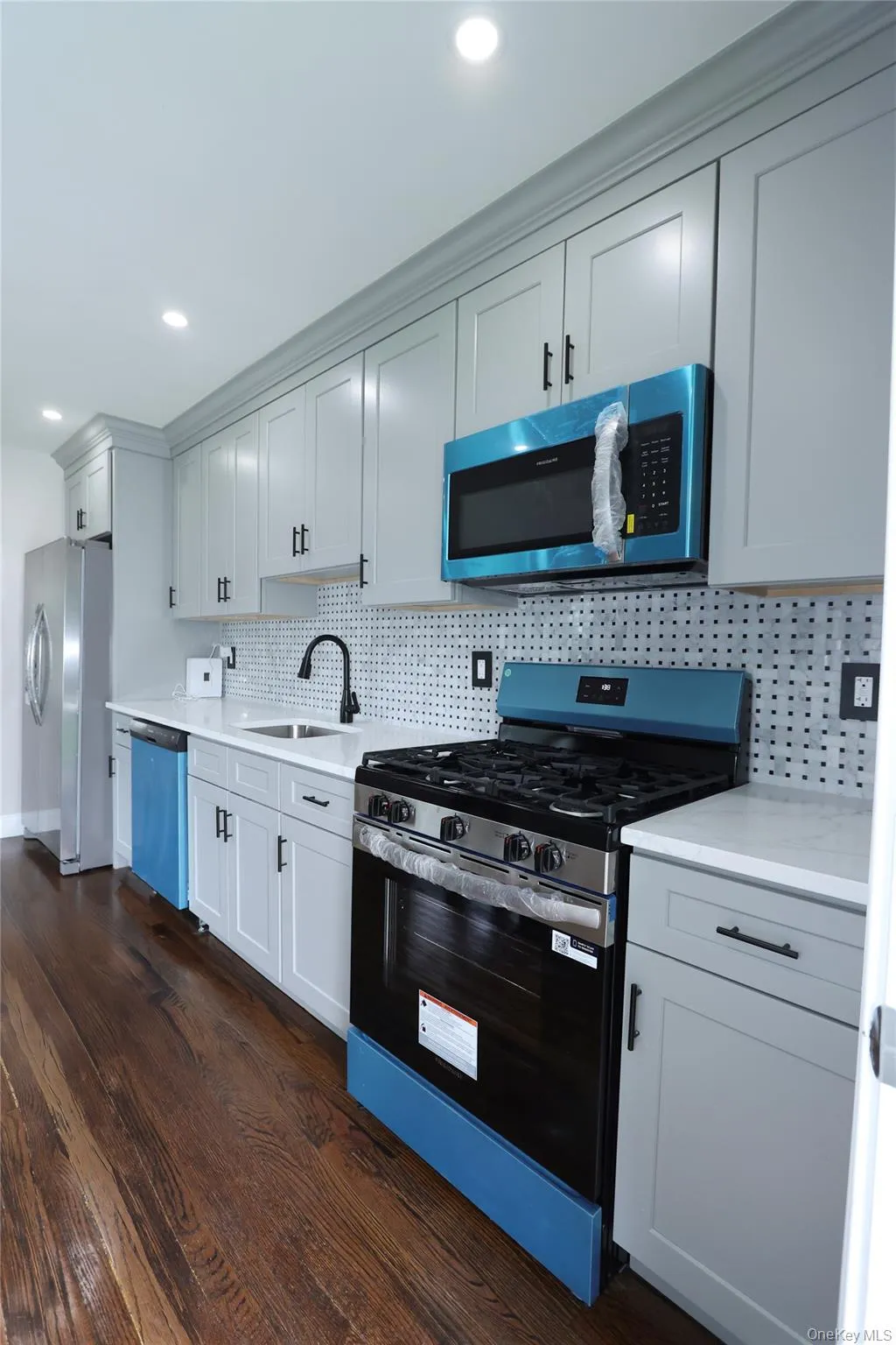 Galley kitchen featuring light gray cabinetry, dark wood-finish flooring, white countertops, tile backsplash, and stainless steel appliances Galley kitchen featuring light gray cabinetry, dark wood-finish flooring, white countertops, tile backsplash, and stainless steel appliances