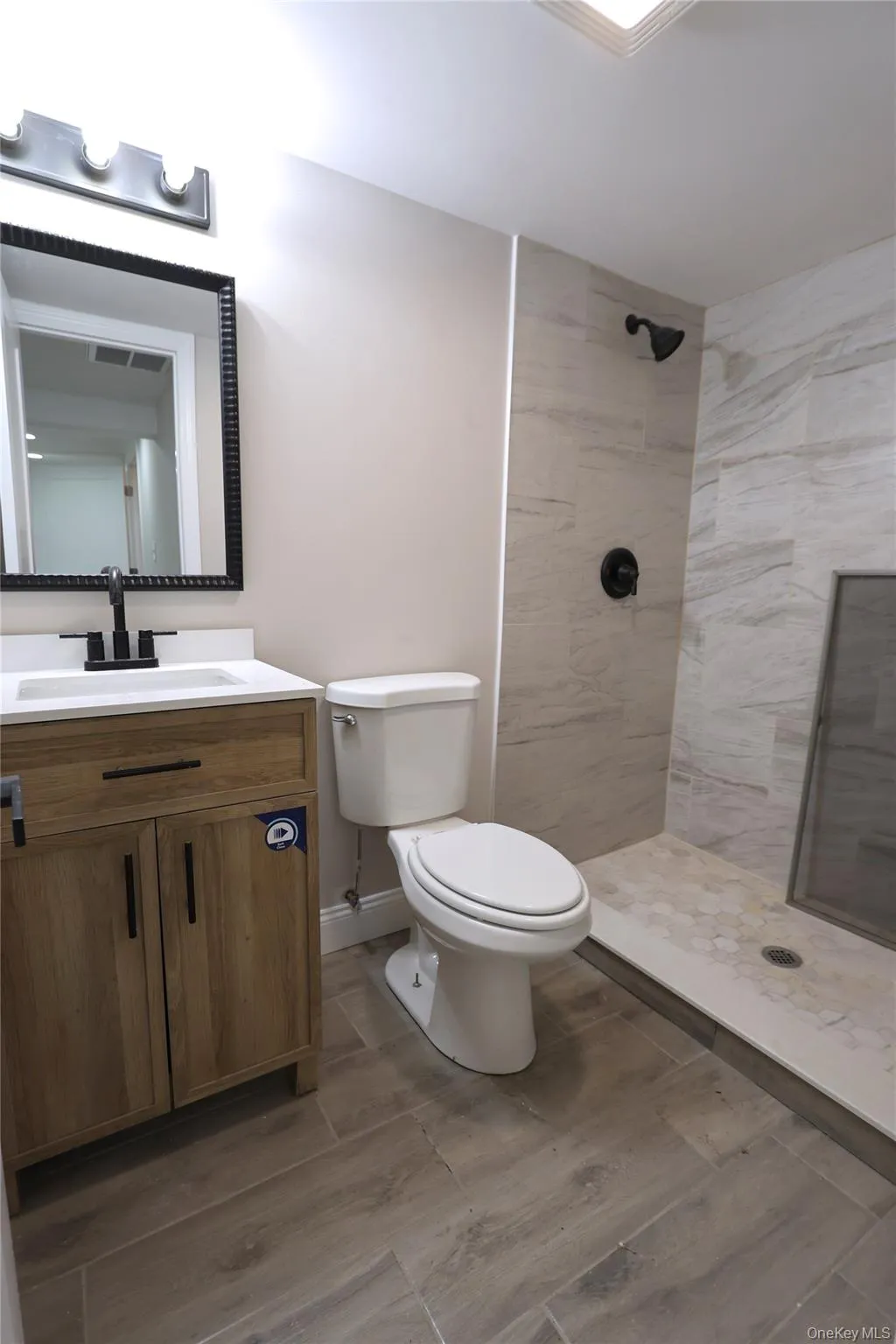 Wood-finish flooring, wood-finish vanity with white countertop, black-framed mirror, and a tiled shower with a dark-finish showerhead Wood-finish flooring, wood-finish vanity with white countertop, black-framed mirror, and a tiled shower with a dark-finish showerhead