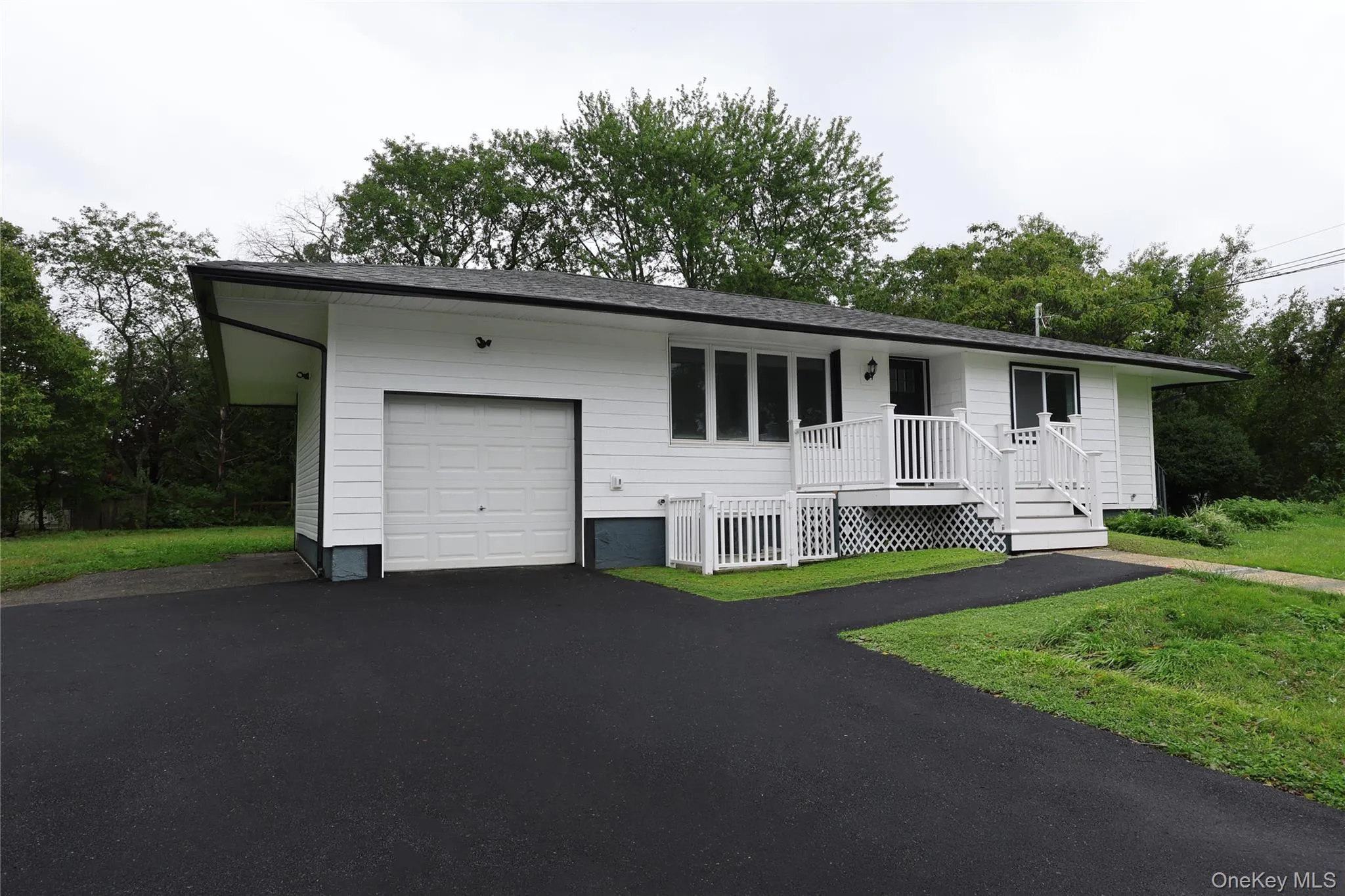 Single-story residence featuring white siding, a dark roof, an attached garage, a front entry deck with white railing, and a paved driveway Single-story residence featuring white siding, a dark roof, an attached garage, a front entry deck with white railing, and a paved driveway