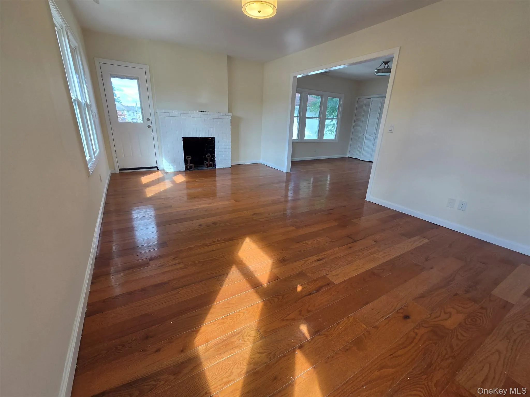 Spacious living area showcasing rich wood-finish flooring throughout, abundant natural light from multiple windows, and a fireplace with white surround Spacious living area showcasing rich wood-finish flooring throughout, abundant natural light from multiple windows, and a fireplace with white surround