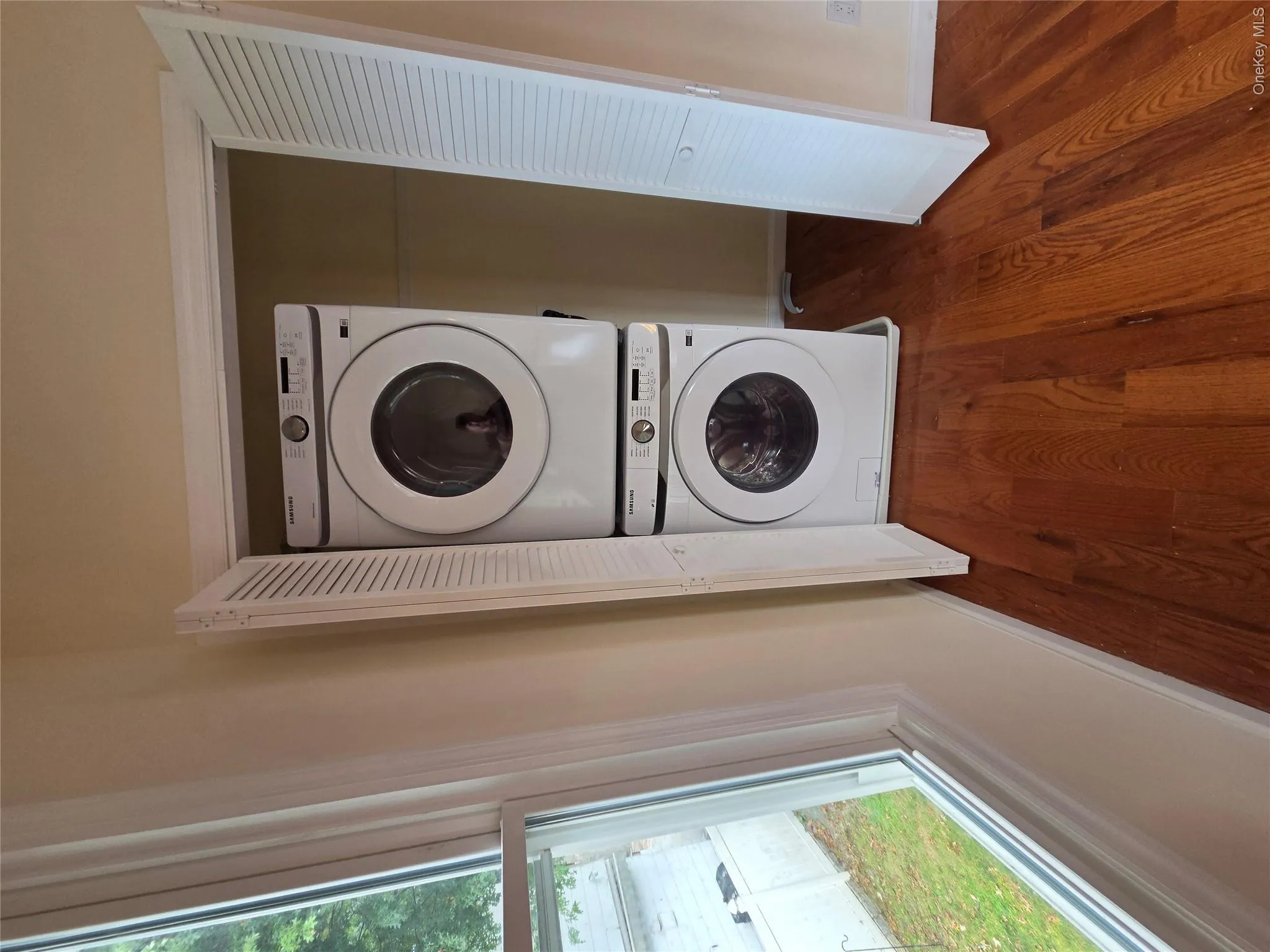 Stackable laundry appliances within a louvered bi-fold door closet, accompanied by a window and wood-finish flooring Stackable laundry appliances within a louvered bi-fold door closet, accompanied by a window and wood-finish flooring