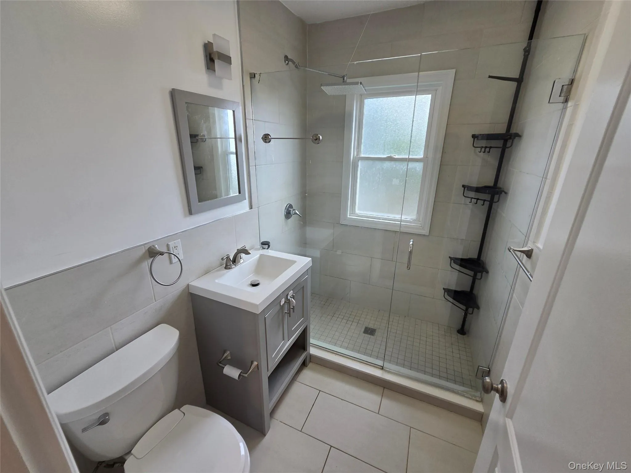 Porcelain tile flooring, glass-enclosed shower, single vanity with integrated sink, contemporary wall sconce, and framed mirror Porcelain tile flooring, glass-enclosed shower, single vanity with integrated sink, contemporary wall sconce, and framed mirror