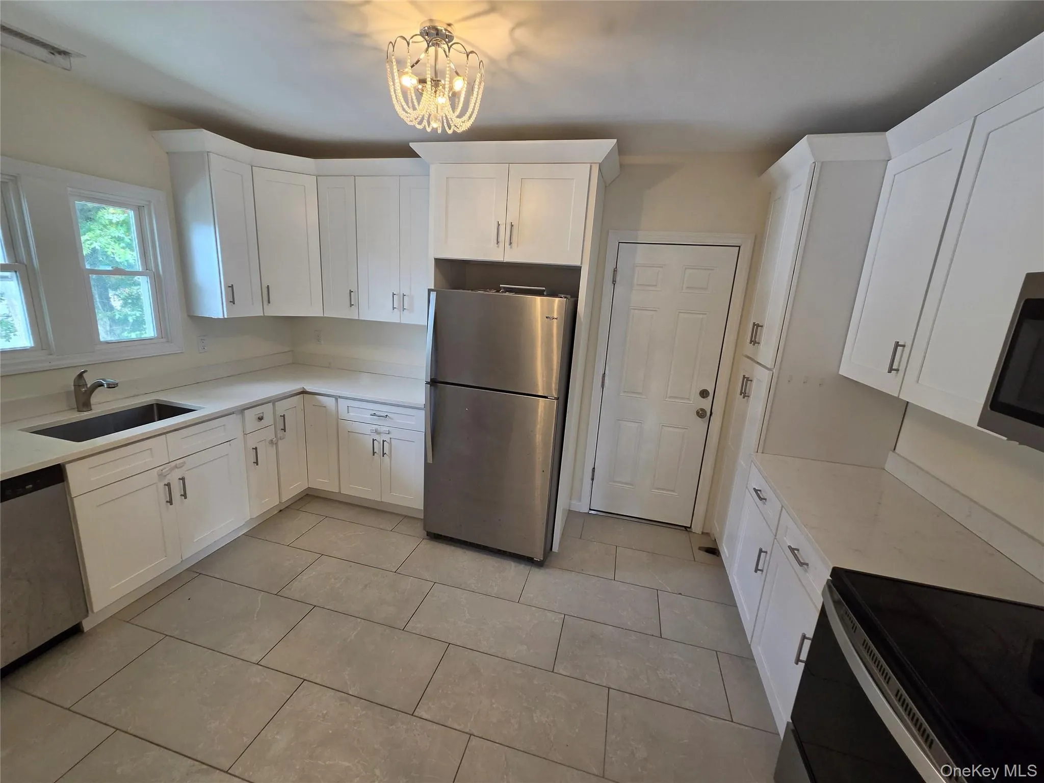 White shaker cabinetry with brushed nickel hardware, light-toned countertops, stainless steel appliances, ceramic tile flooring, and a decorative ceiling light fixture White shaker cabinetry with brushed nickel hardware, light-toned countertops, stainless steel appliances, ceramic tile flooring, and a decorative ceiling light fixture