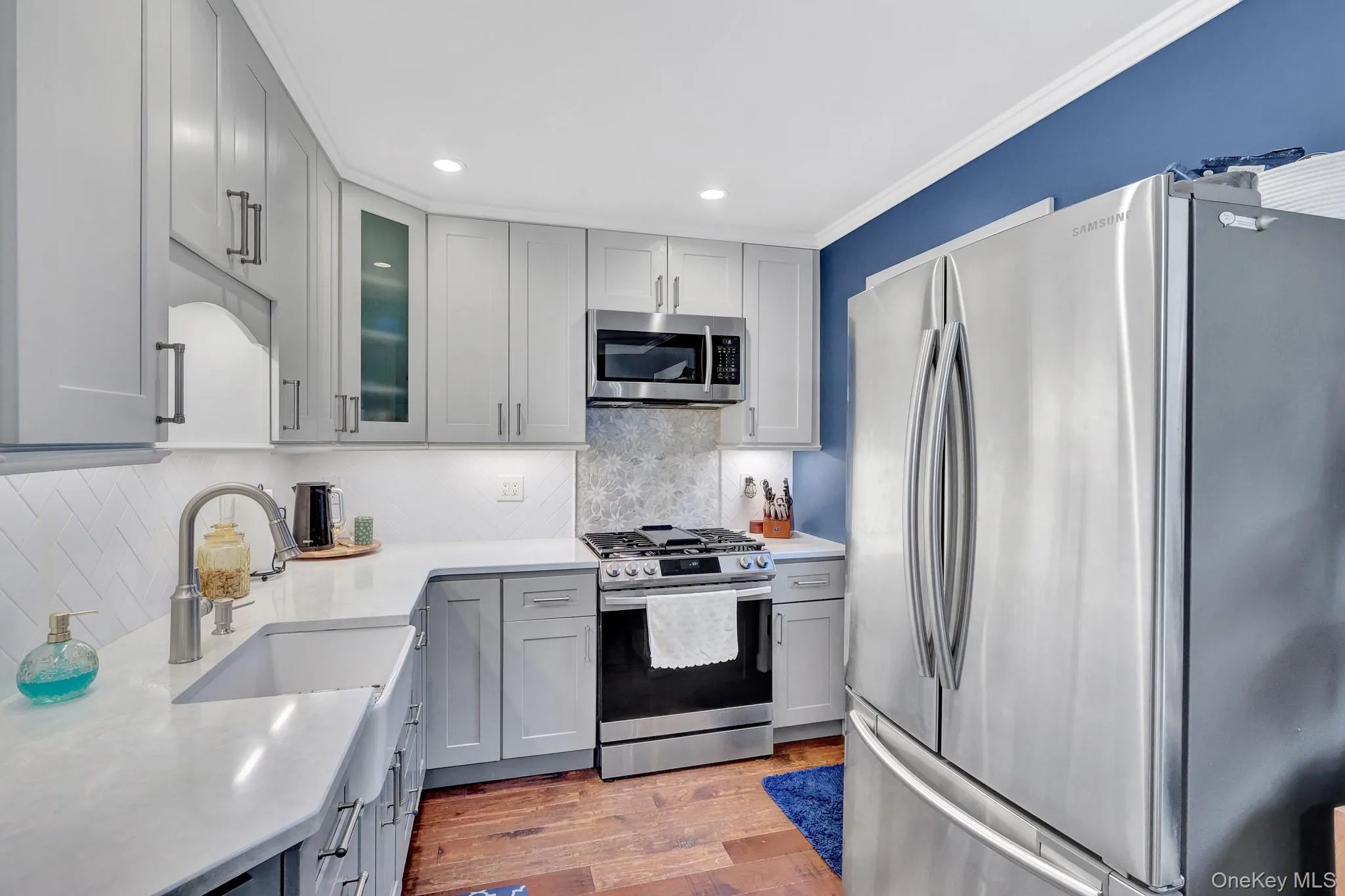 Light gray shaker cabinetry, stainless steel appliances, a farmhouse sink, white countertops, and wood-finish flooring Light gray shaker cabinetry, stainless steel appliances, a farmhouse sink, white countertops, and wood-finish flooring