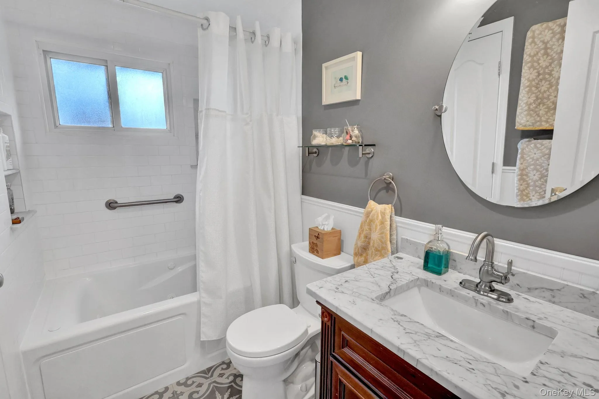 Bathroom featuring a wood-finish vanity with an integrated sink and stone countertop, a circular wall-mounted mirror, and a built-in bathtub with subway tile surround and grab bar Bathroom featuring a wood-finish vanity with an integrated sink and stone countertop, a circular wall-mounted mirror, and a built-in bathtub with subway tile surround and grab bar