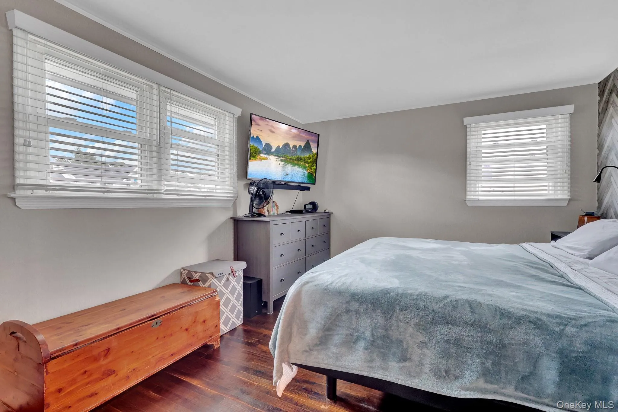 Bright secondary bedroom featuring multiple windows with plantation shutters, wood-finish flooring, and soft neutral wall tones Bright secondary bedroom featuring multiple windows with plantation shutters, wood-finish flooring, and soft neutral wall tones
