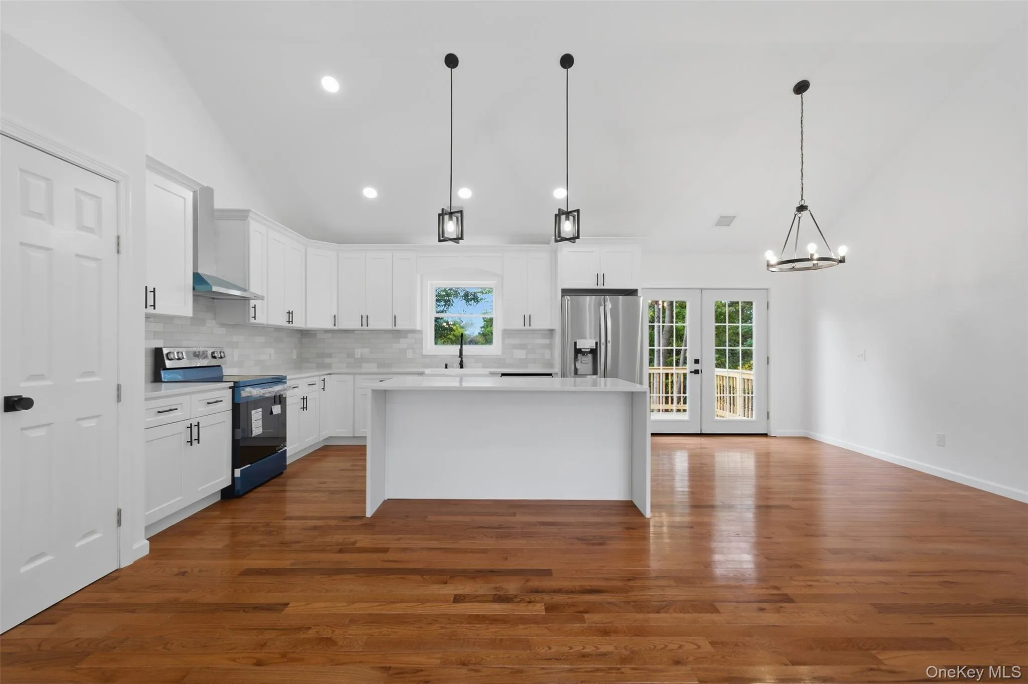 High ceiling with recessed lighting, wood-finish flooring, white kitchen cabinetry with stainless steel appliances, kitchen island with countertop, and double doors opening to an exterior deck High ceiling with recessed lighting, wood-finish flooring, white kitchen cabinetry with stainless steel appliances, kitchen island with countertop, and double doors opening to an exterior deck
