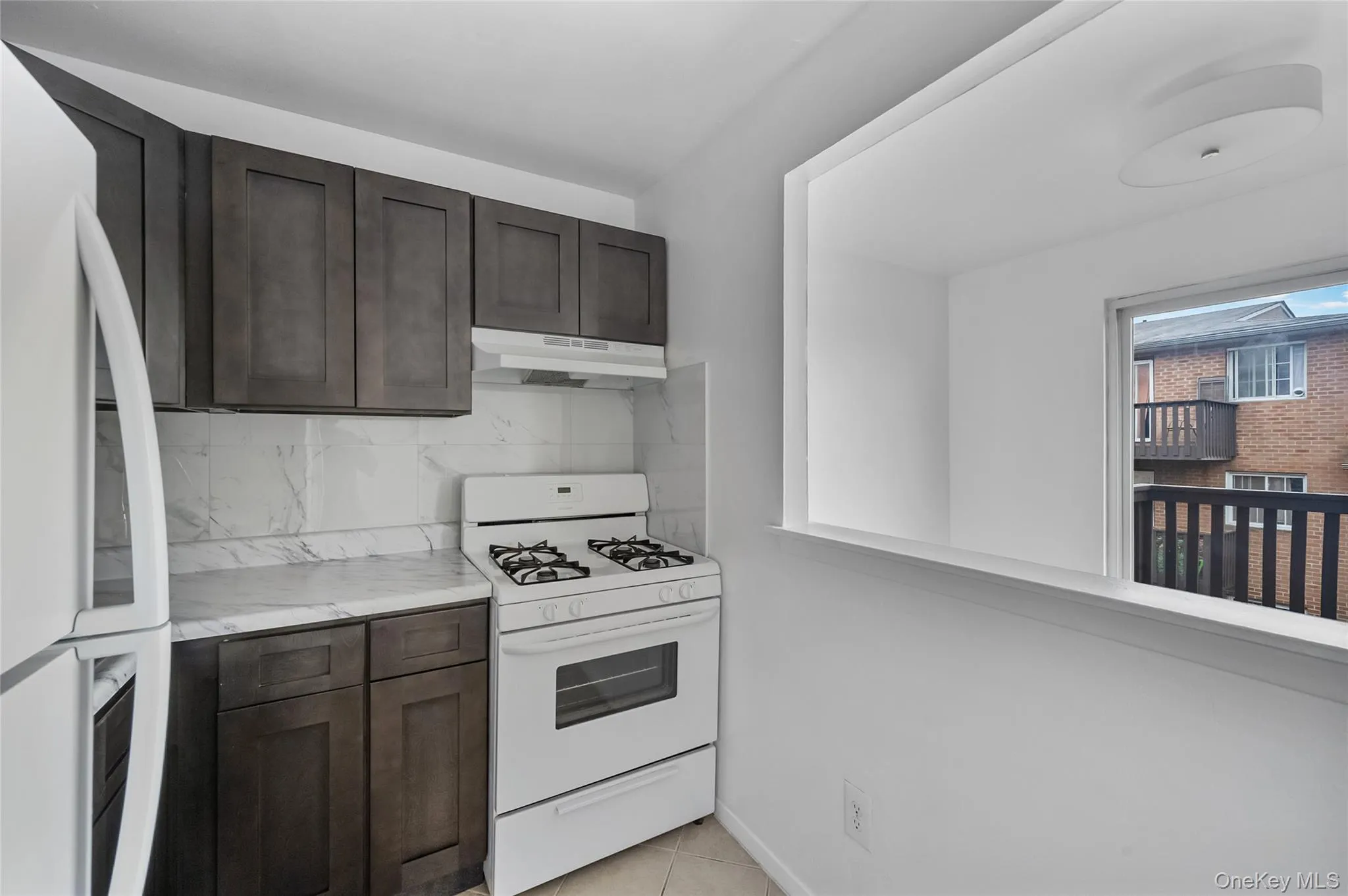 Kitchen featuring dark brown cabinets, white appliances, backsplash, under cabinet range hood, and light tile patterned flooring Kitchen featuring dark brown cabinets, white appliances, backsplash, under cabinet range hood, and light tile patterned flooring