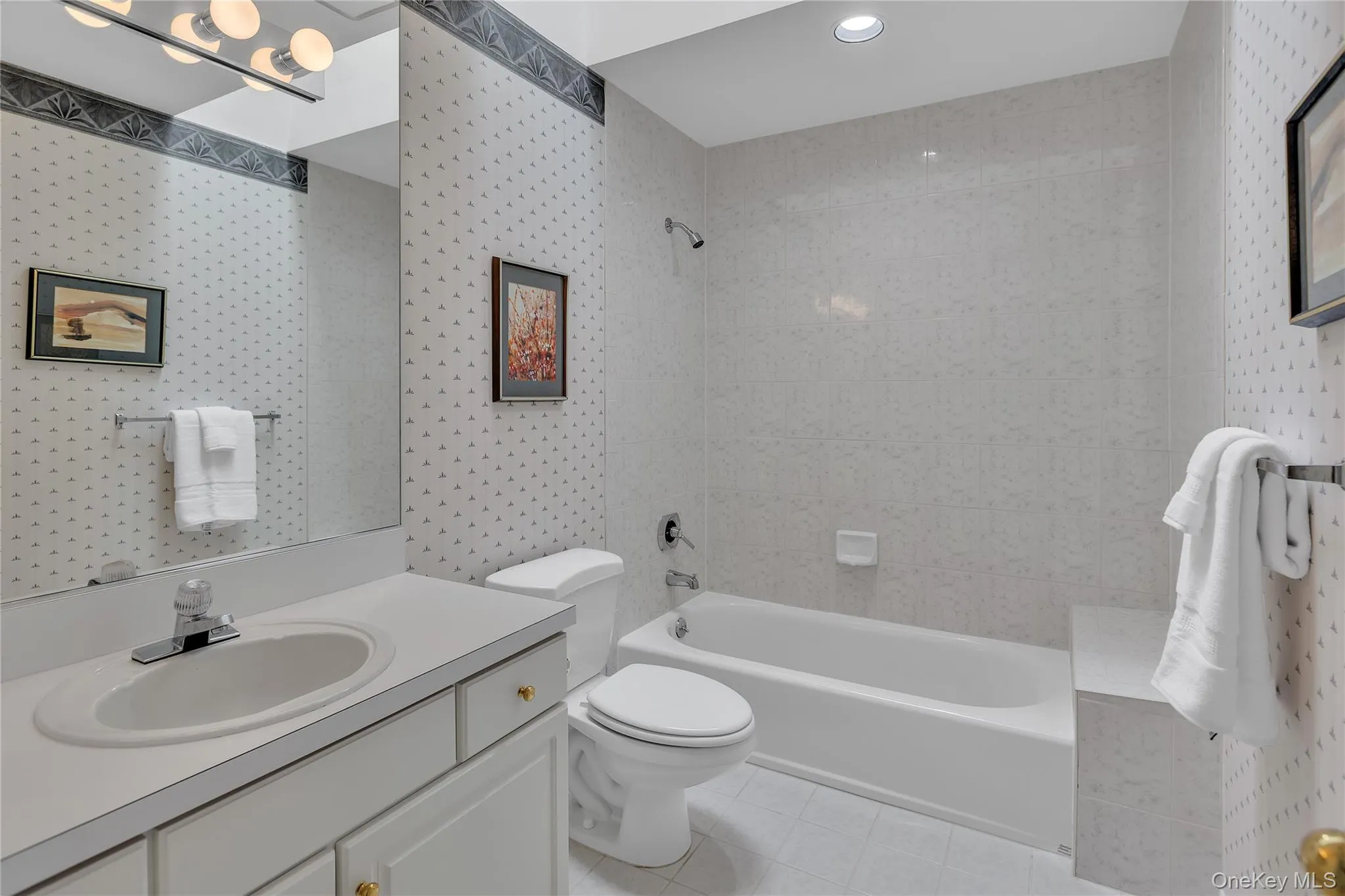 Full bathroom featuring a single vanity with an integrated sink, a large mirror, a chrome multi-bulb fixture, a toilet, and a shower and tub combined with white tile surround and a skylight. Full bathroom featuring a single vanity with an integrated sink, a large mirror, a chrome multi-bulb fixture, a toilet, and a shower and tub combined with white tile surround and a skylight.