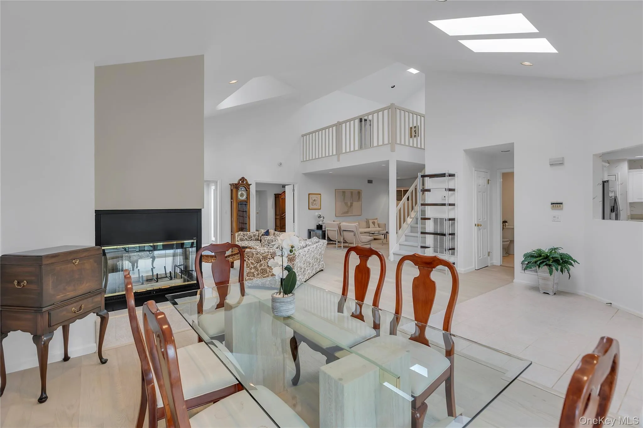 Dining area featuring a glass tabletop, a black-framed fireplace, vaulted ceilings, skylights and a partial view of a staircase with a wood railing. Dining area featuring a glass tabletop, a black-framed fireplace, vaulted ceilings, skylights and a partial view of a staircase with a wood railing.