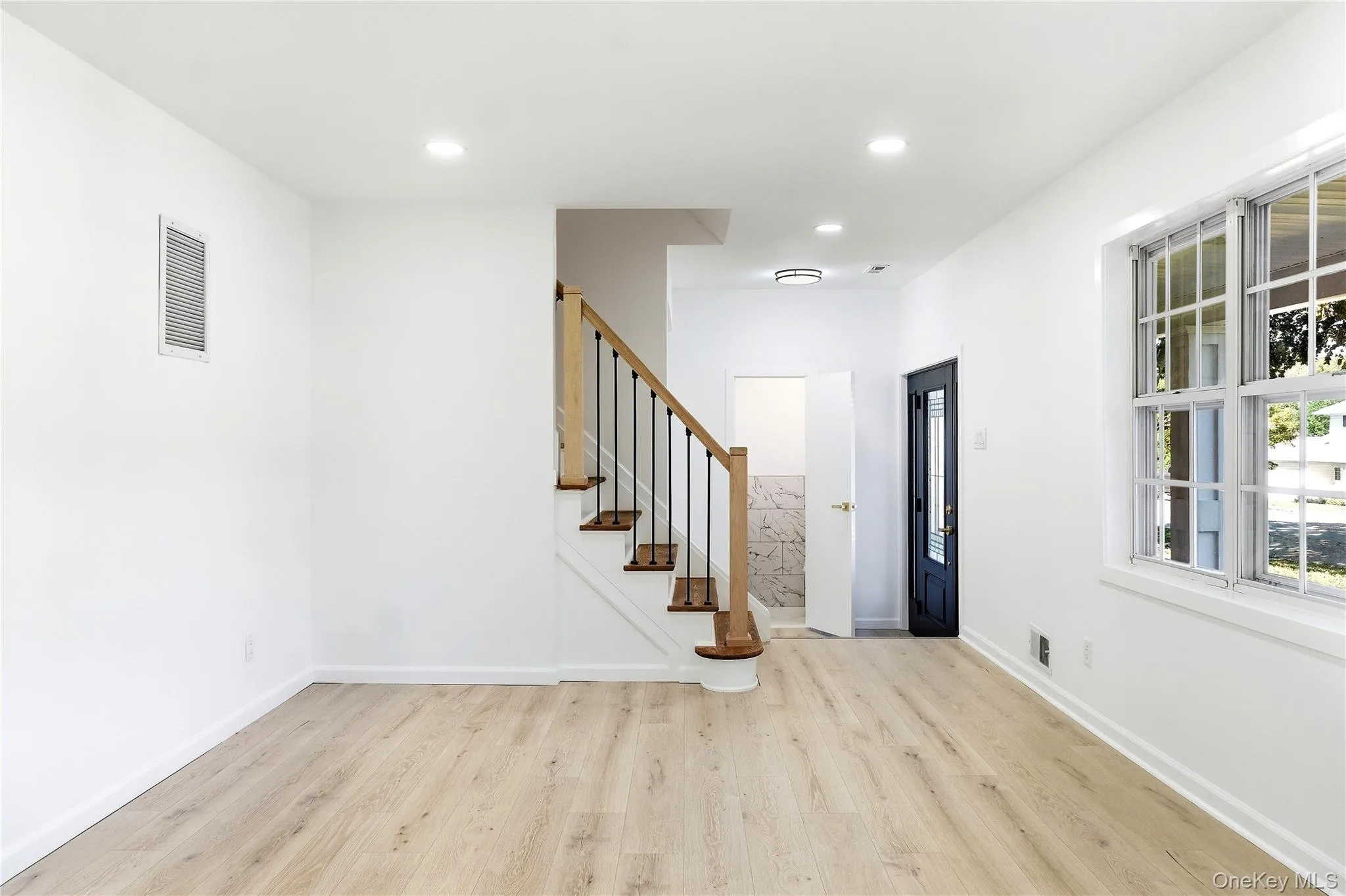 Light wood-finish flooring, white walls, recessed ceiling lighting, and a staircase with wood risers and a black metal railing Light wood-finish flooring, white walls, recessed ceiling lighting, and a staircase with wood risers and a black metal railing