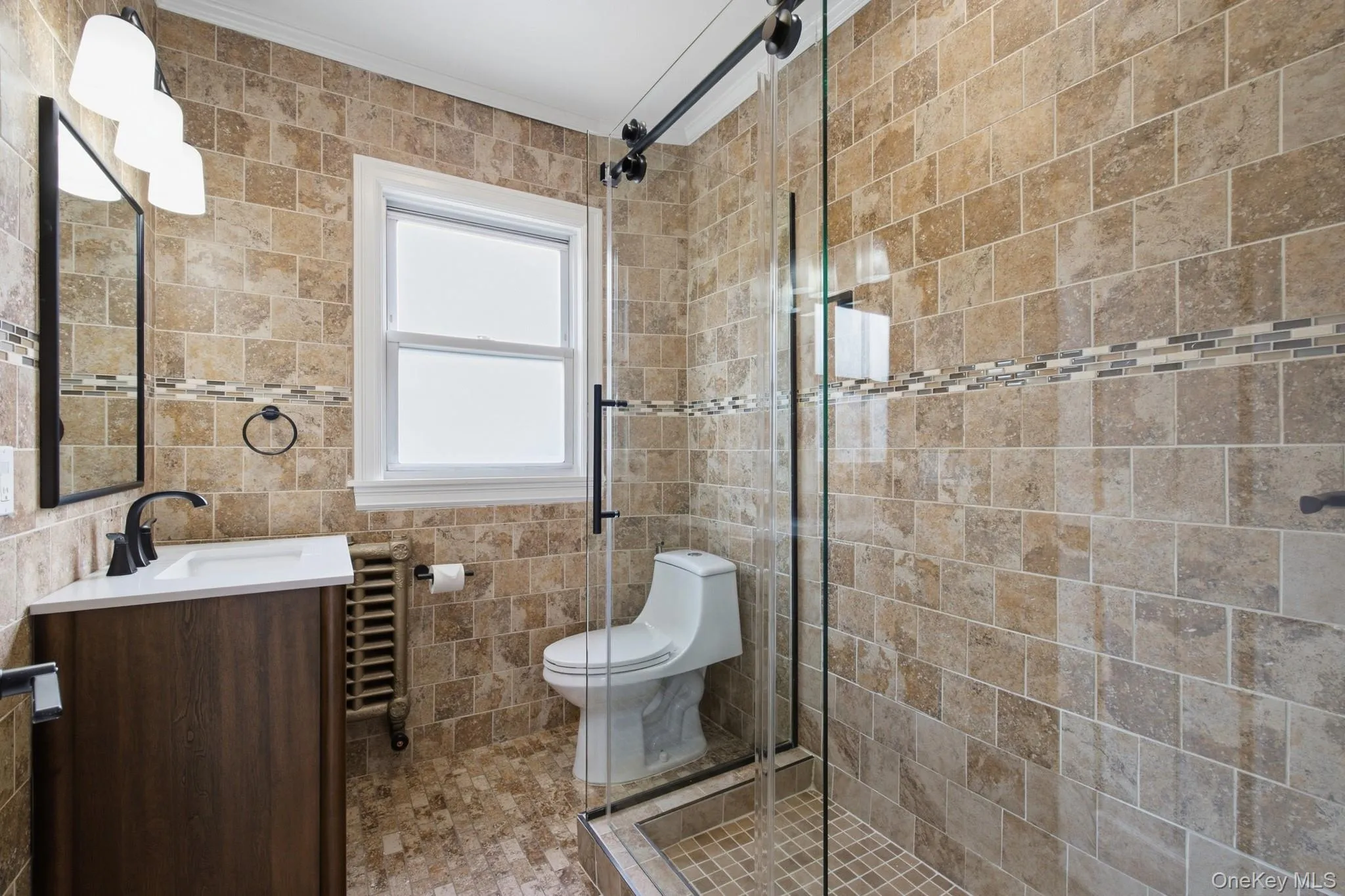 Bathroom featuring floor-to-ceiling natural stone wall and floor tile, glass-enclosed shower with matte black hardware, single vanity with integrated sink, and a modern light fixture Bathroom featuring floor-to-ceiling natural stone wall and floor tile, glass-enclosed shower with matte black hardware, single vanity with integrated sink, and a modern light fixture
