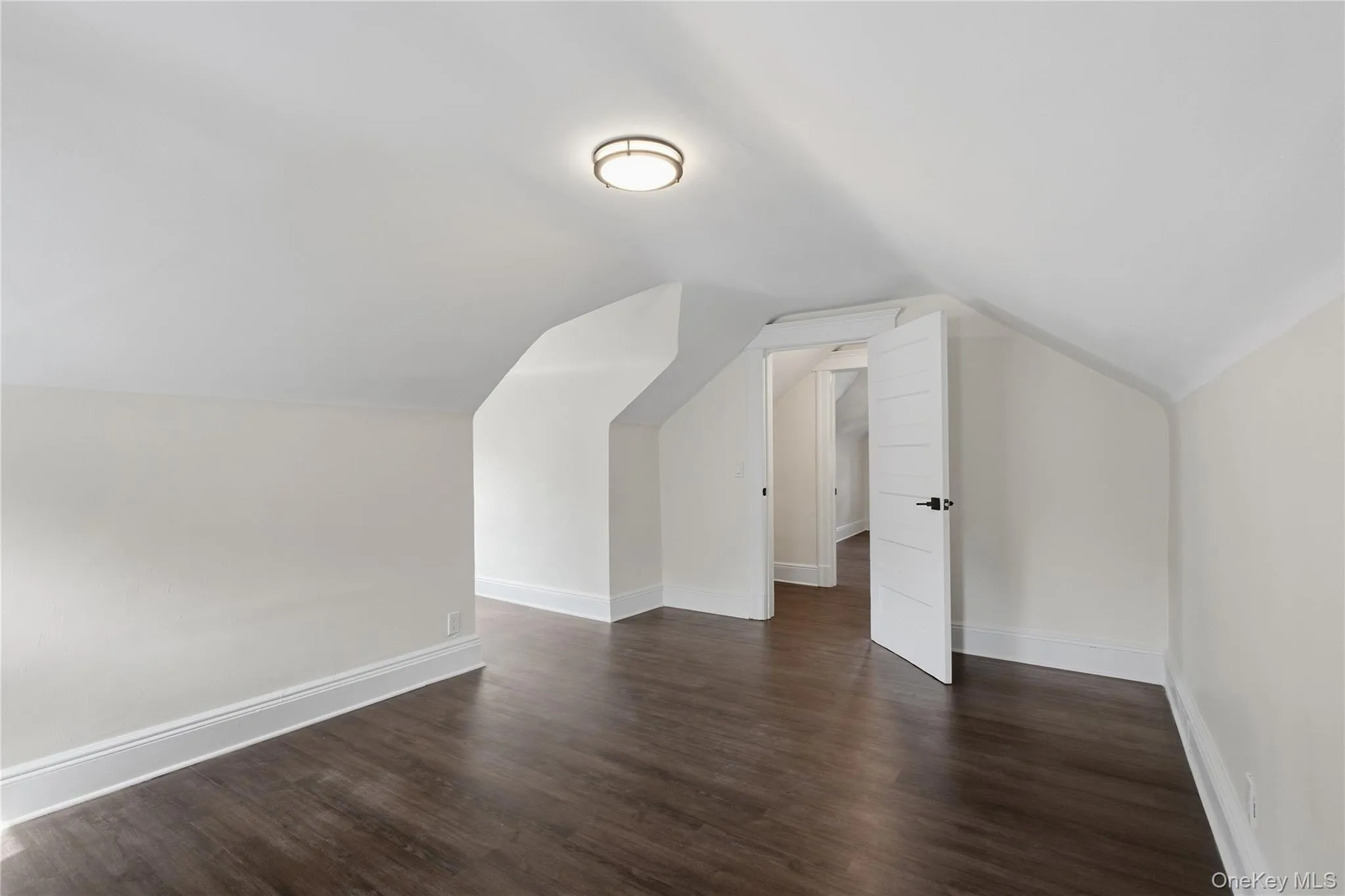 Sloped ceilings, wood-finish flooring, white trim, and a flush-mount ceiling light Sloped ceilings, wood-finish flooring, white trim, and a flush-mount ceiling light
