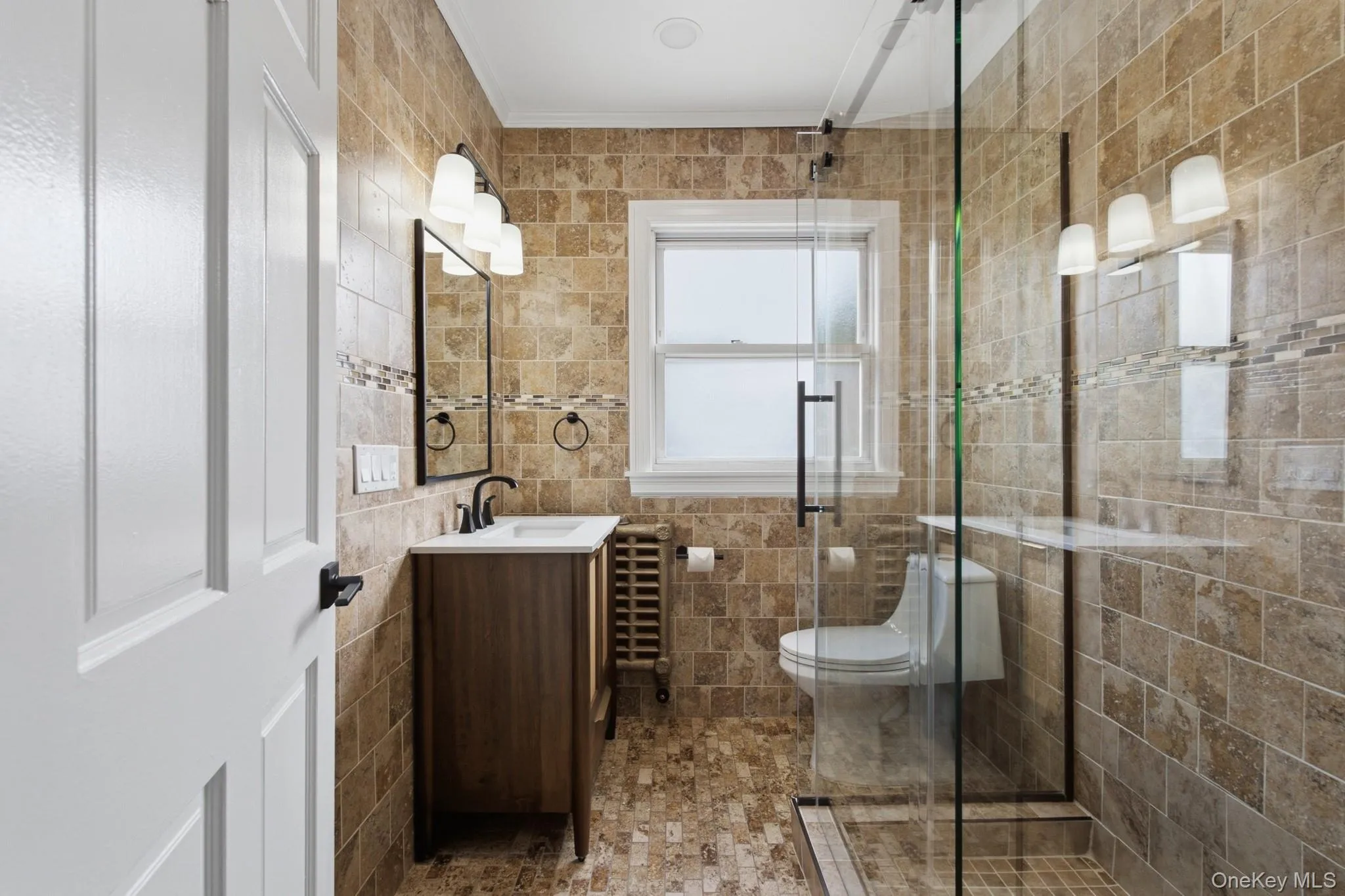Tiled bathroom featuring a glass-enclosed shower, wood-finish vanity, and a window Tiled bathroom featuring a glass-enclosed shower, wood-finish vanity, and a window