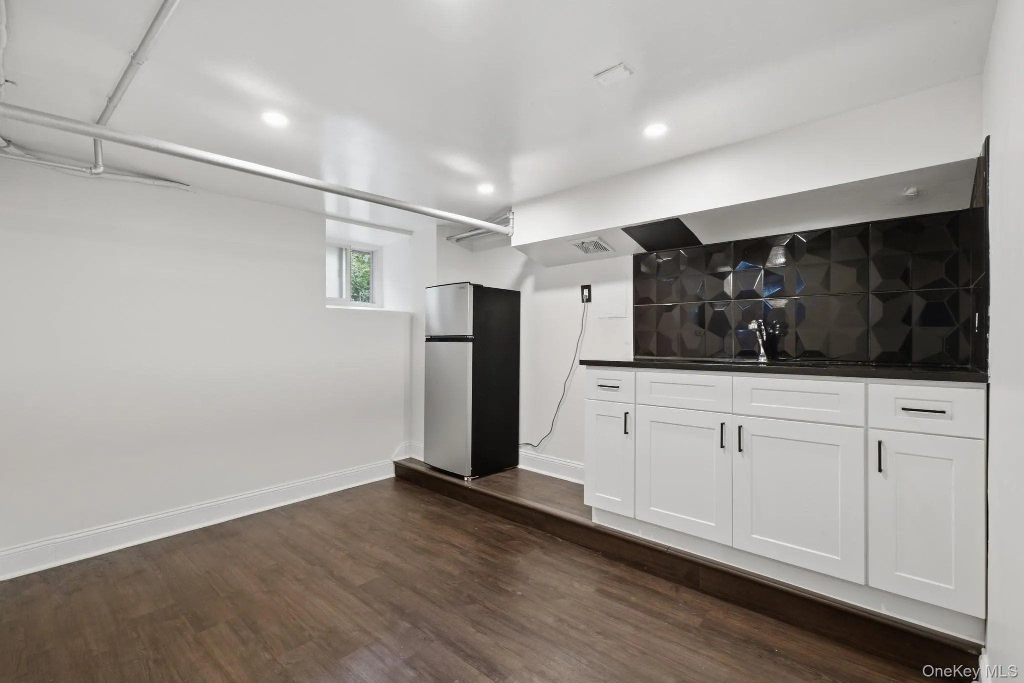 Wood-finish flooring, recessed lighting, white cabinetry with dark hardware, dark countertop, and geometric tile backsplash Wood-finish flooring, recessed lighting, white cabinetry with dark hardware, dark countertop, and geometric tile backsplash