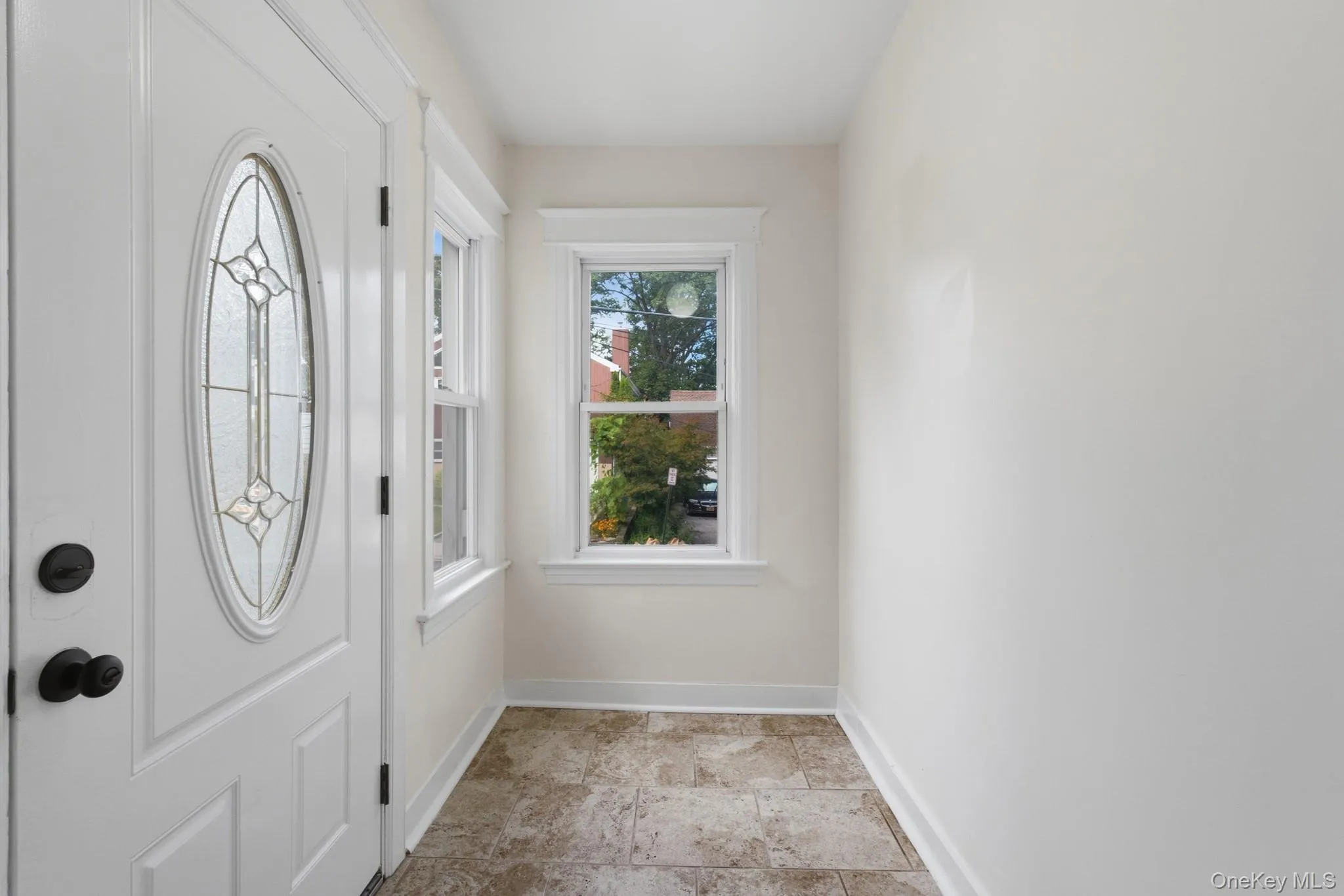 Entryway with a white door featuring an oval leaded glass inset, two double-hung windows with white trim, and tiled flooring Entryway with a white door featuring an oval leaded glass inset, two double-hung windows with white trim, and tiled flooring