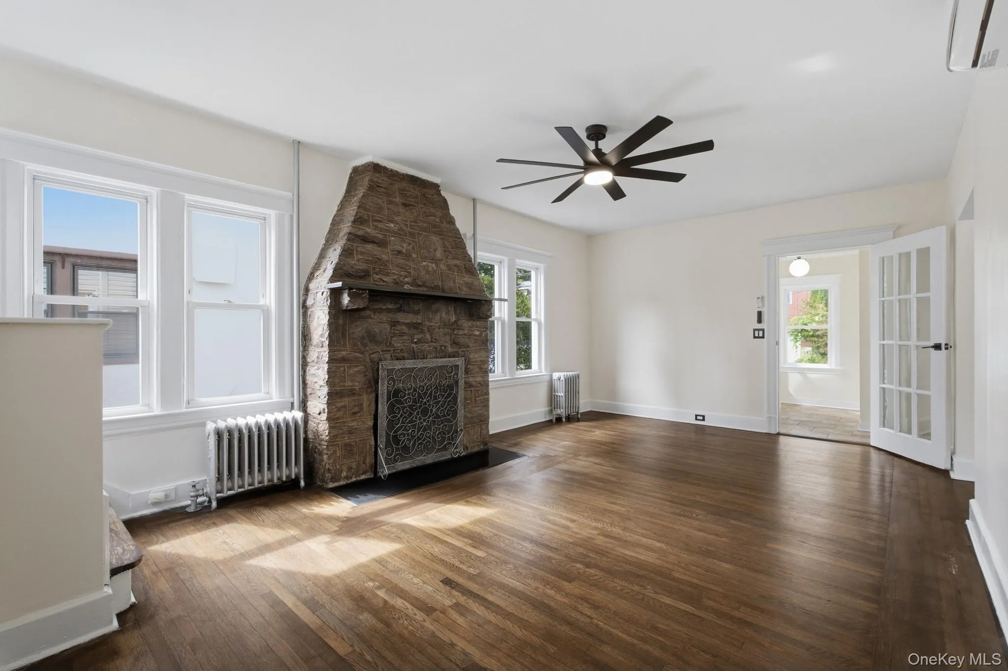 Spacious room featuring wood-finish flooring, a stone fireplace, multiple windows, and a ceiling fan with integrated lighting Spacious room featuring wood-finish flooring, a stone fireplace, multiple windows, and a ceiling fan with integrated lighting