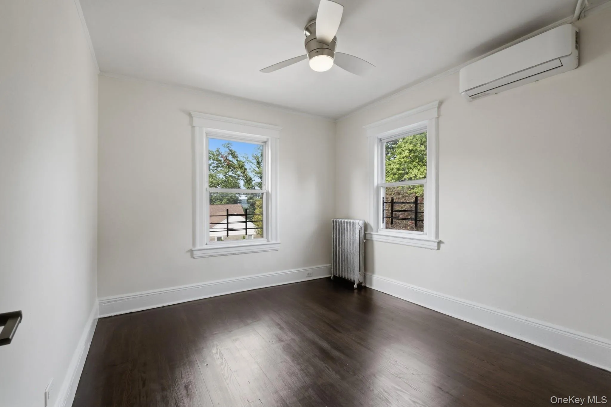 Dark wood-finish flooring, white walls, two windows with white trim, a ceiling fan with integrated lighting, and a wall-mounted air conditioning unit Dark wood-finish flooring, white walls, two windows with white trim, a ceiling fan with integrated lighting, and a wall-mounted air conditioning unit