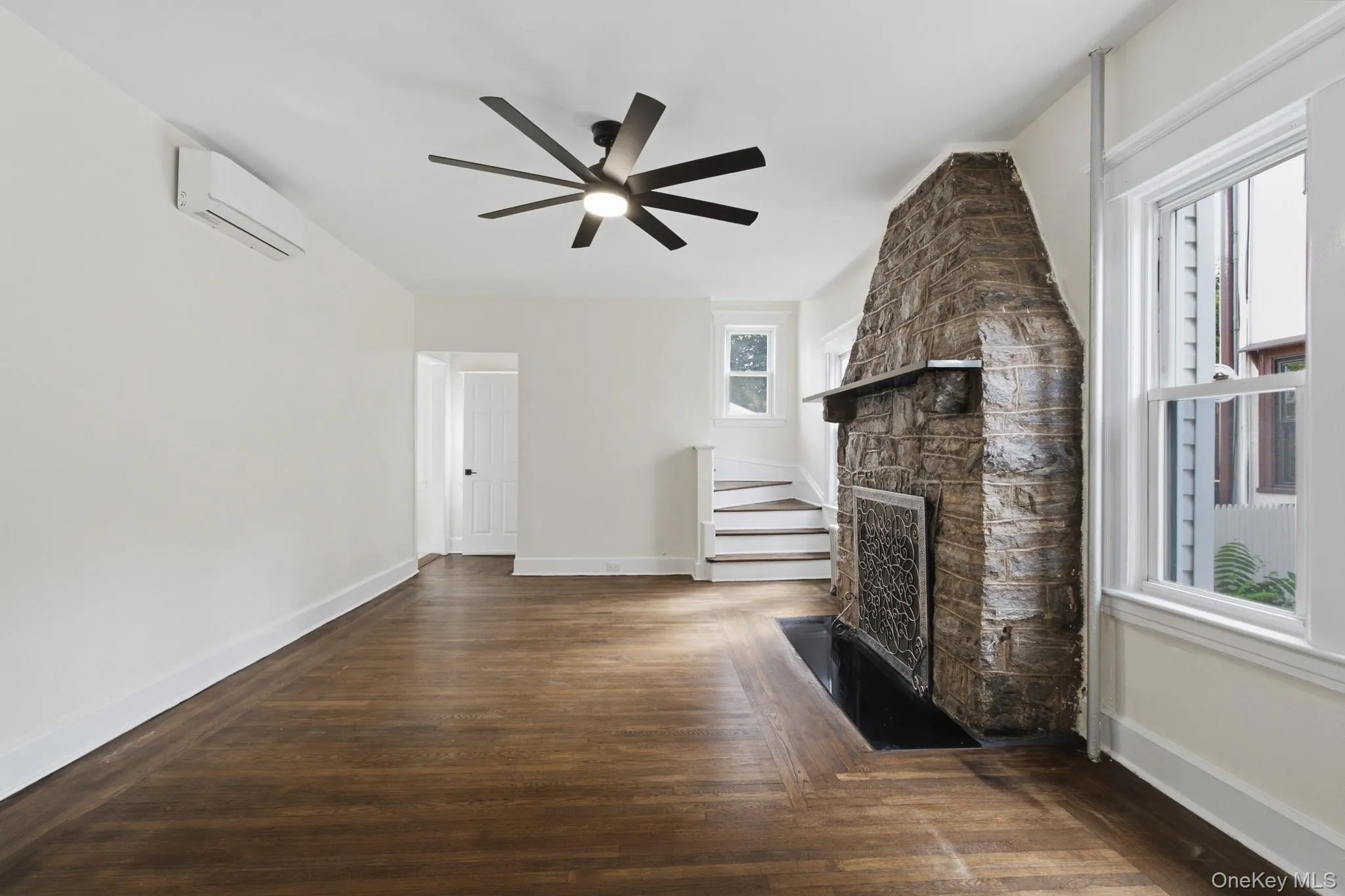 Hardwood-finish flooring, stone fireplace with hearth, ceiling fan, split-system air conditioning unit, and white trim Hardwood-finish flooring, stone fireplace with hearth, ceiling fan, split-system air conditioning unit, and white trim