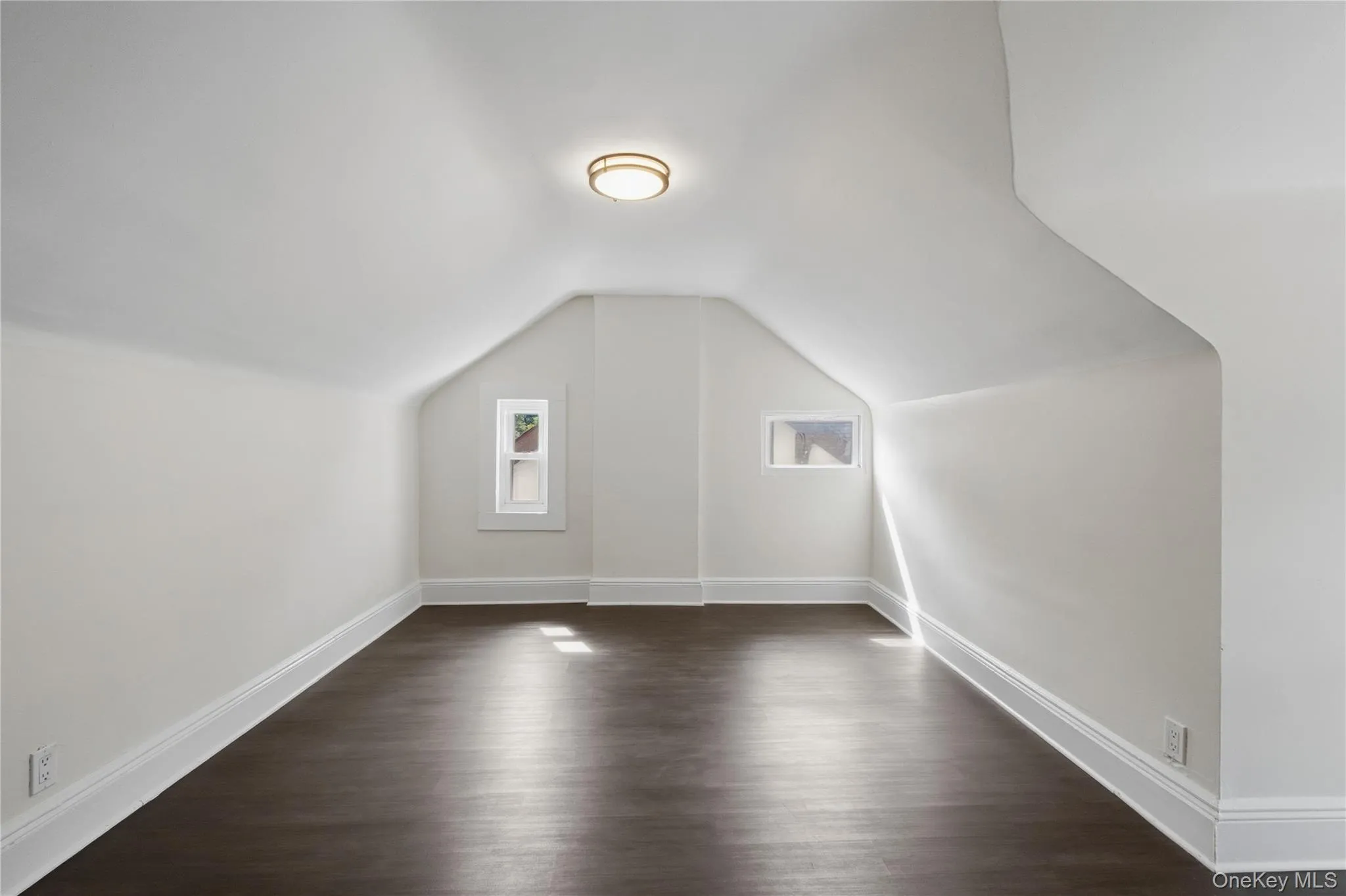 Sloped ceilings, two distinct windows, wood-finish flooring, and a flush-mount ceiling light fixture Sloped ceilings, two distinct windows, wood-finish flooring, and a flush-mount ceiling light fixture