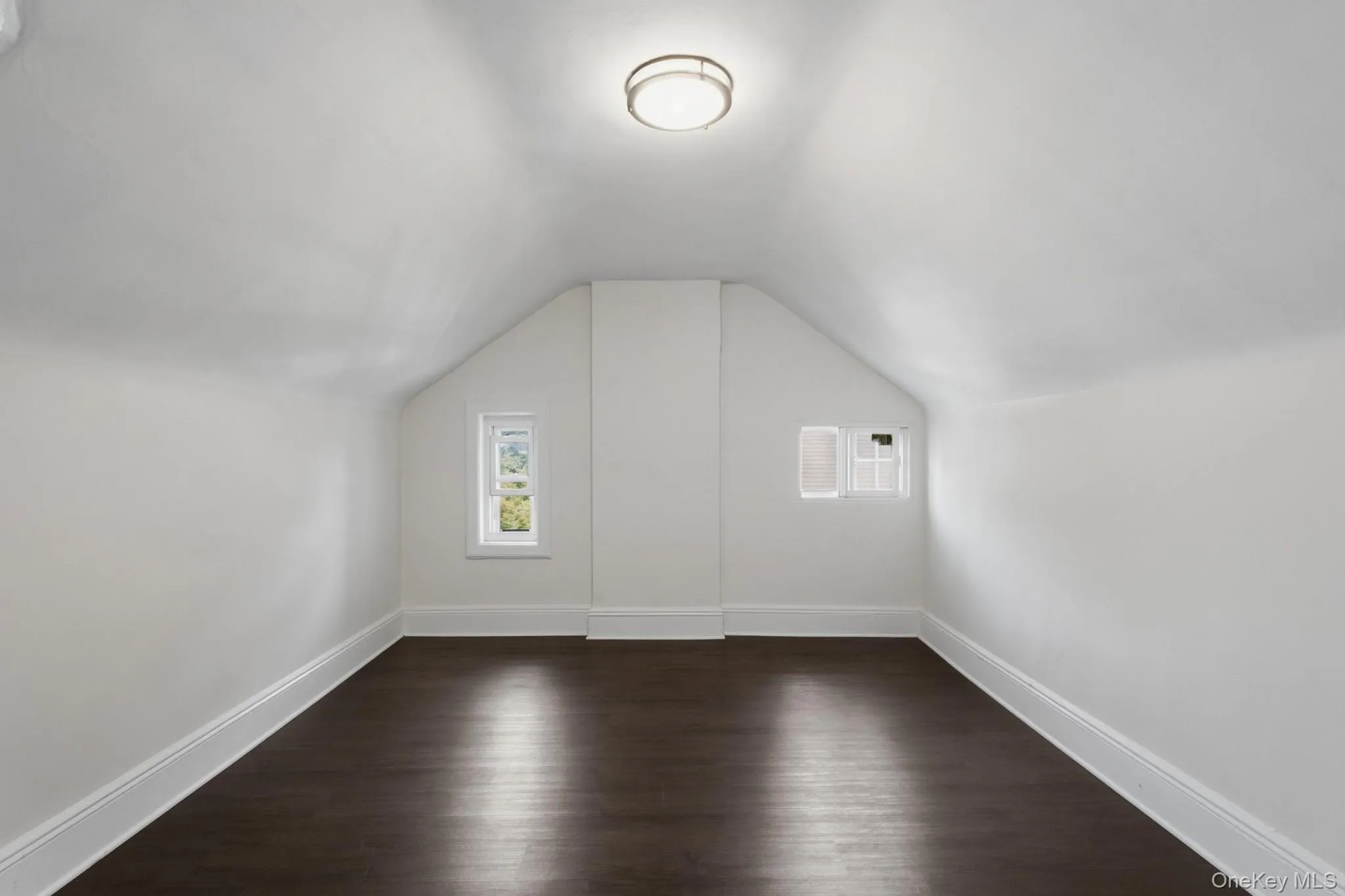 Sloped ceiling, dark wood-finish flooring, white walls, flush mount ceiling light, and two windows Sloped ceiling, dark wood-finish flooring, white walls, flush mount ceiling light, and two windows