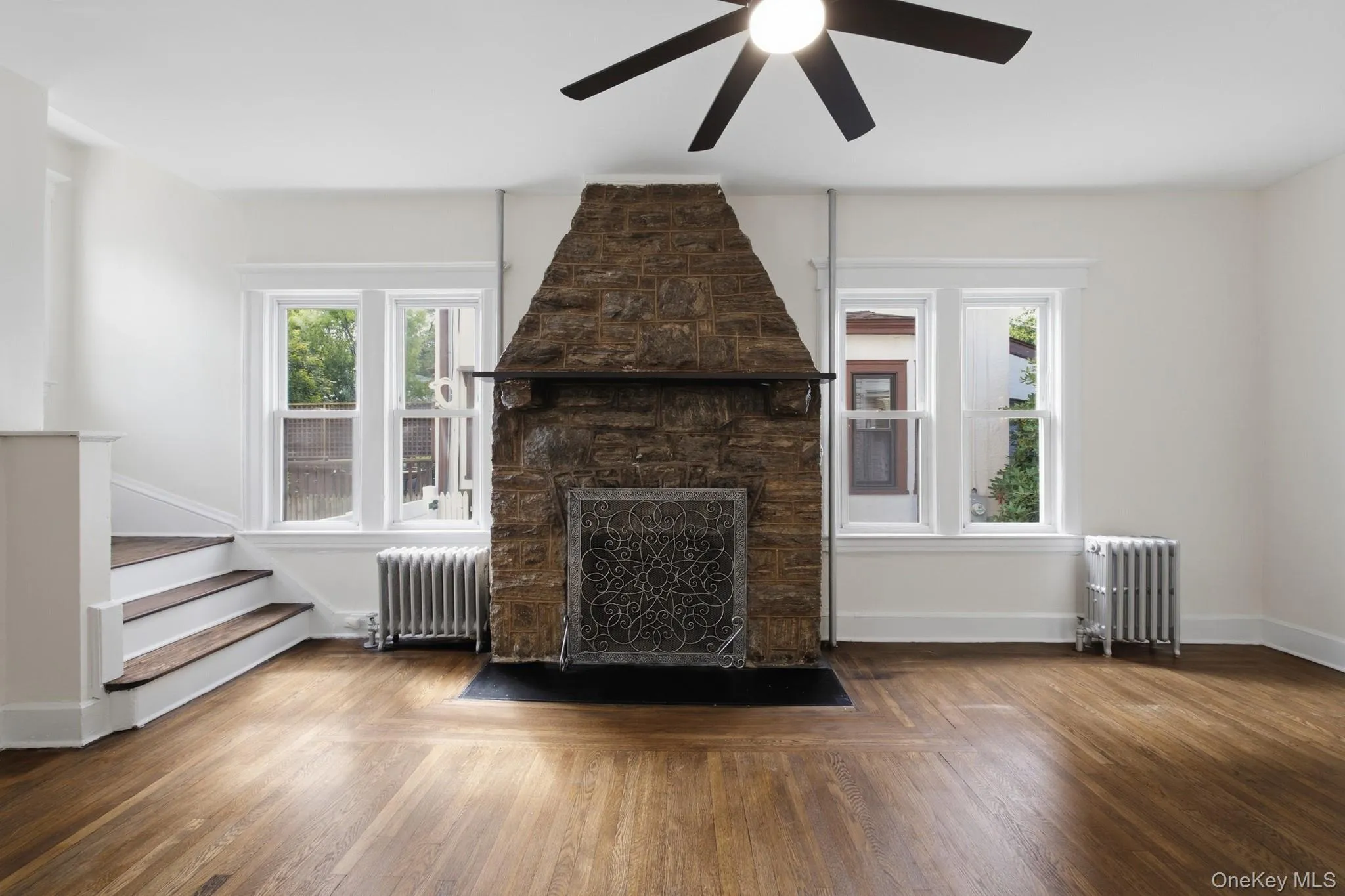 Hardwood flooring, stone-clad fireplace with mantel, ceiling fan with integrated lighting, dual sash windows, and white painted walls Hardwood flooring, stone-clad fireplace with mantel, ceiling fan with integrated lighting, dual sash windows, and white painted walls