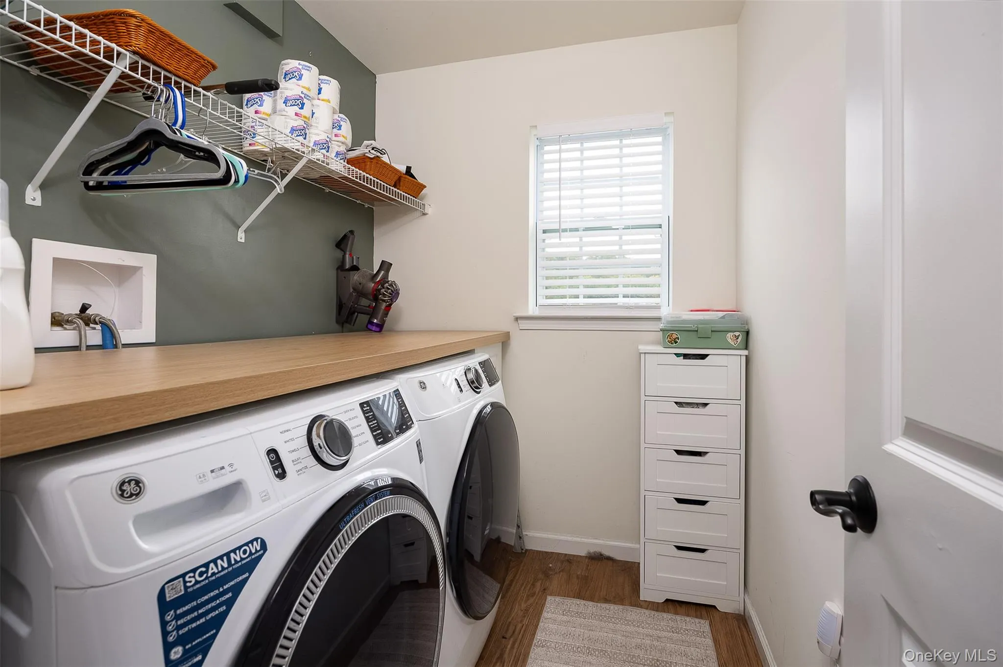Laundry room with light wood-finish countertop, window with blinds, wire shelving, and a white paneled door with black hardware Laundry room with light wood-finish countertop, window with blinds, wire shelving, and a white paneled door with black hardware