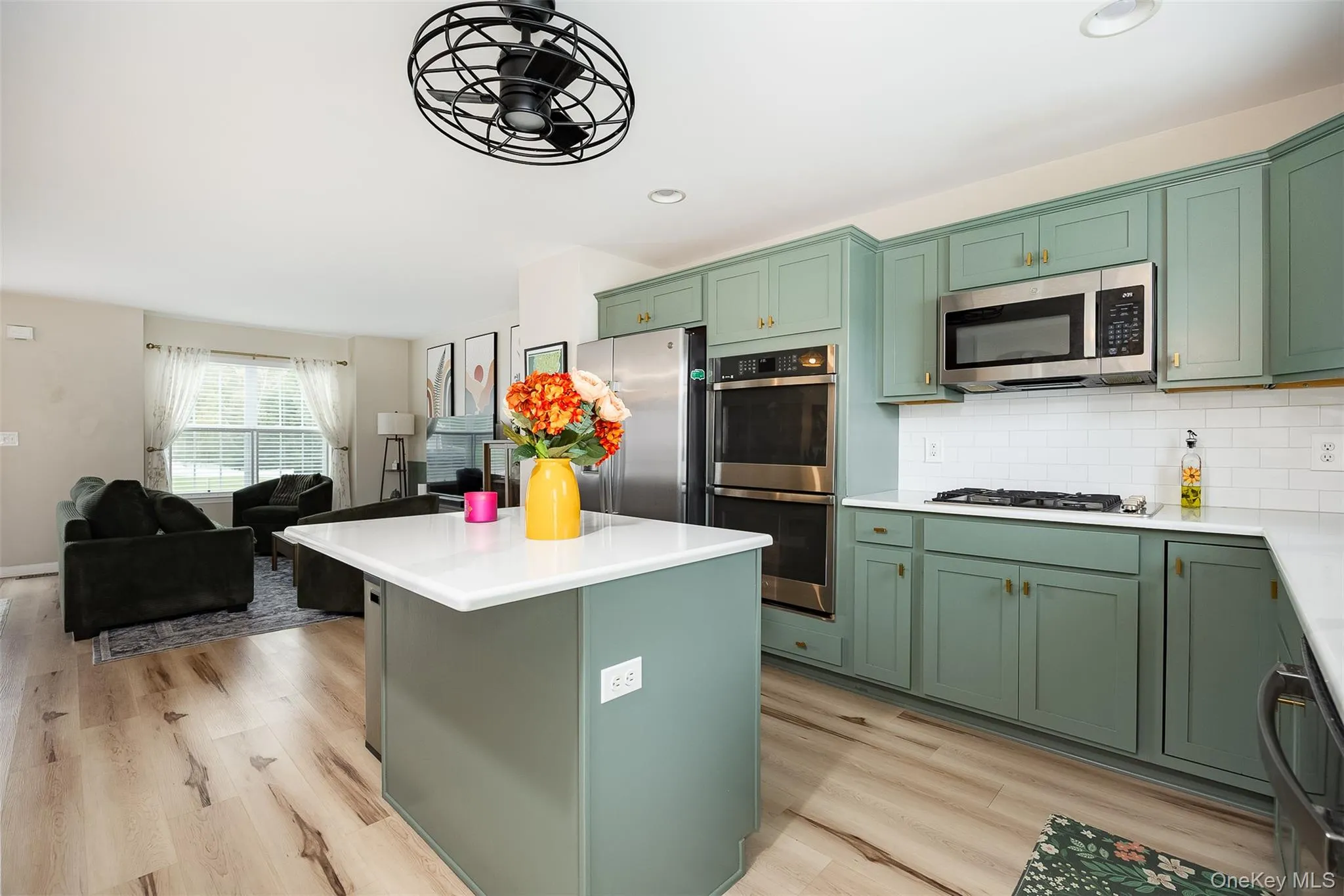 Open-concept kitchen with sage green cabinetry, stainless steel appliances, white subway tile backsplash, and a central island with a white countertop Open-concept kitchen with sage green cabinetry, stainless steel appliances, white subway tile backsplash, and a central island with a white countertop