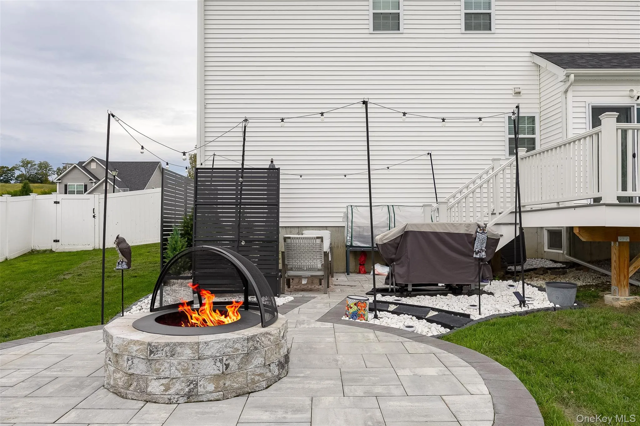 Stone-clad fire pit, paver patio, string lighting, white vinyl fencing, and white siding Stone-clad fire pit, paver patio, string lighting, white vinyl fencing, and white siding