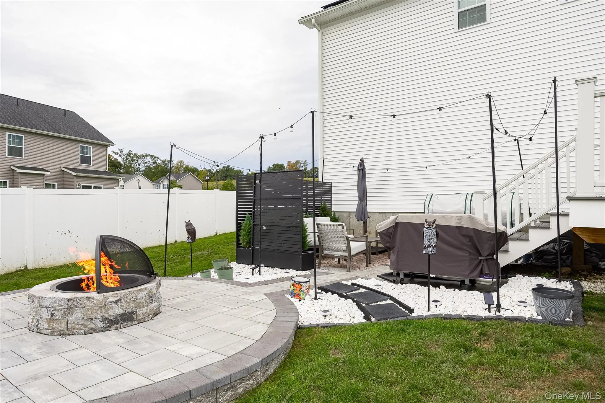 Paver patio with a stone fire pit, string lights, and a white privacy fence Paver patio with a stone fire pit, string lights, and a white privacy fence