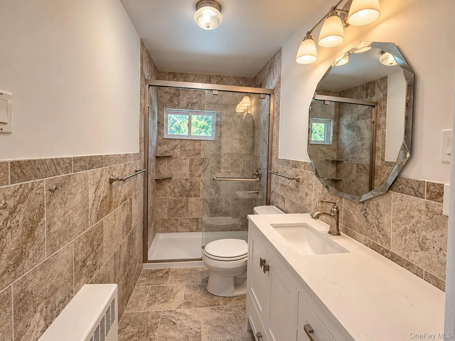 Tiled flooring and wainscoting, glass-enclosed shower with window, white vanity with integrated sink, contemporary vanity lighting, and decorative mirror Tiled flooring and wainscoting, glass-enclosed shower with window, white vanity with integrated sink, contemporary vanity lighting, and decorative mirror