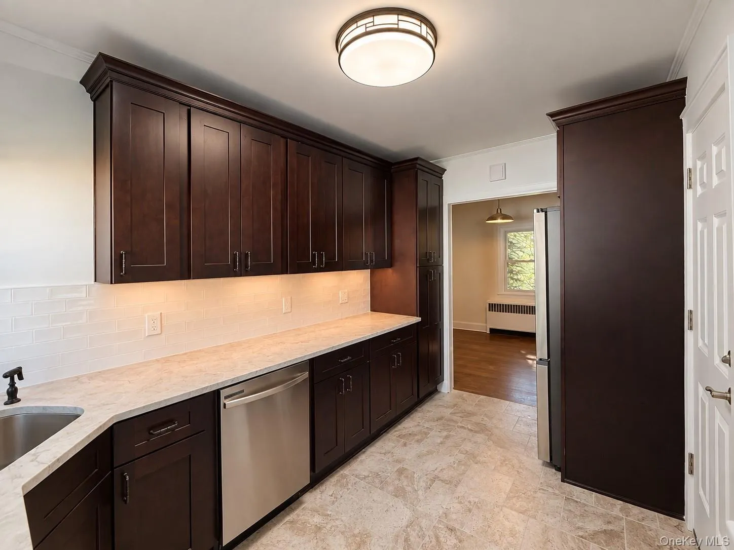 Dark wood cabinetry with crown molding, light-toned countertop, stainless steel dishwasher, tile flooring, and white subway tile backsplash Dark wood cabinetry with crown molding, light-toned countertop, stainless steel dishwasher, tile flooring, and white subway tile backsplash