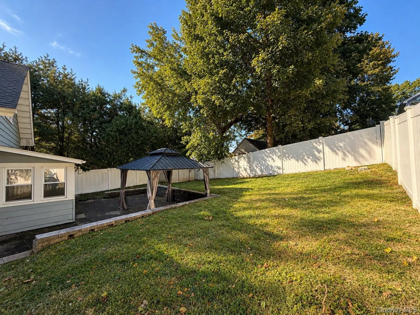 Expansive backyard featuring a gazebo with a dark-finish roof, a stone-block retaining wall, a paved patio area, and a white privacy fence Expansive backyard featuring a gazebo with a dark-finish roof, a stone-block retaining wall, a paved patio area, and a white privacy fence