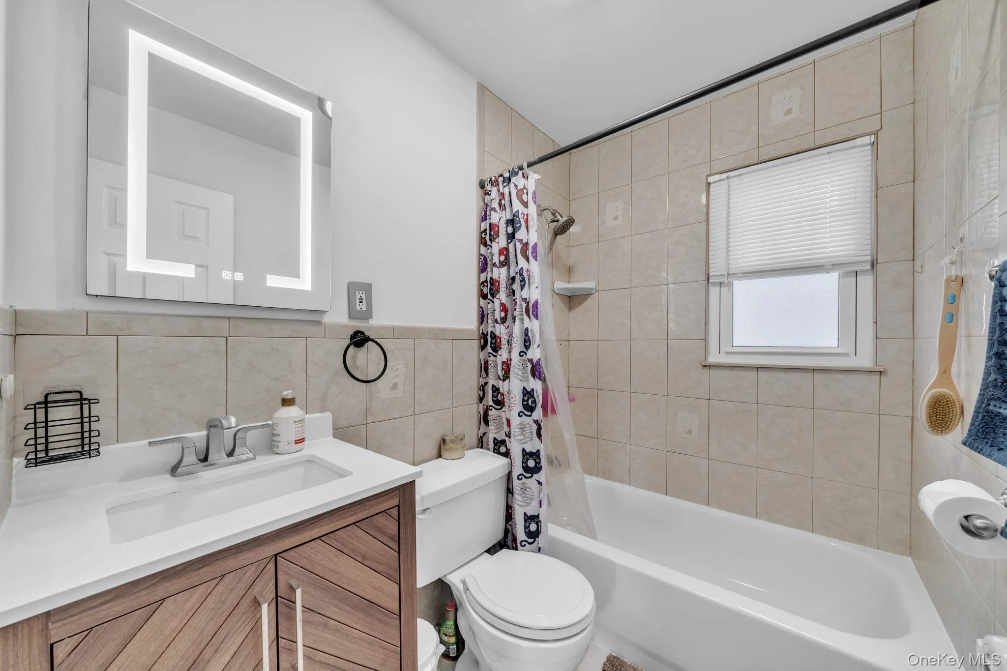 Illuminated vanity mirror, wood-finish vanity with an integrated sink, full bathtub with a shower fixture, and tiled walls Illuminated vanity mirror, wood-finish vanity with an integrated sink, full bathtub with a shower fixture, and tiled walls
