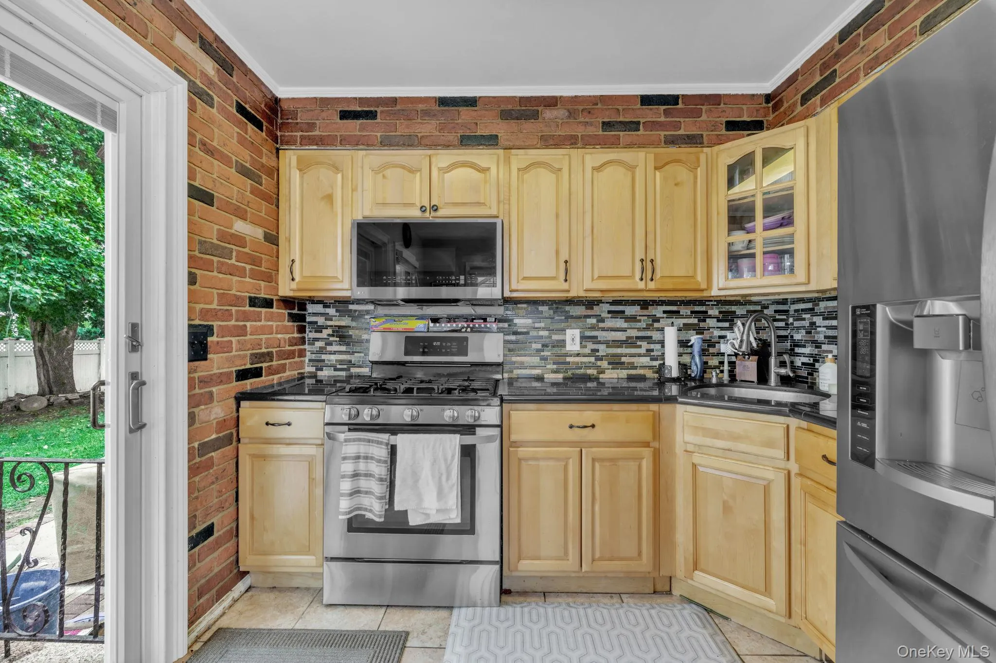 Kitchen featuring light wood cabinetry, a mosaic tile backsplash, black countertops, stainless steel appliances, and brick-patterned wall accents Kitchen featuring light wood cabinetry, a mosaic tile backsplash, black countertops, stainless steel appliances, and brick-patterned wall accents