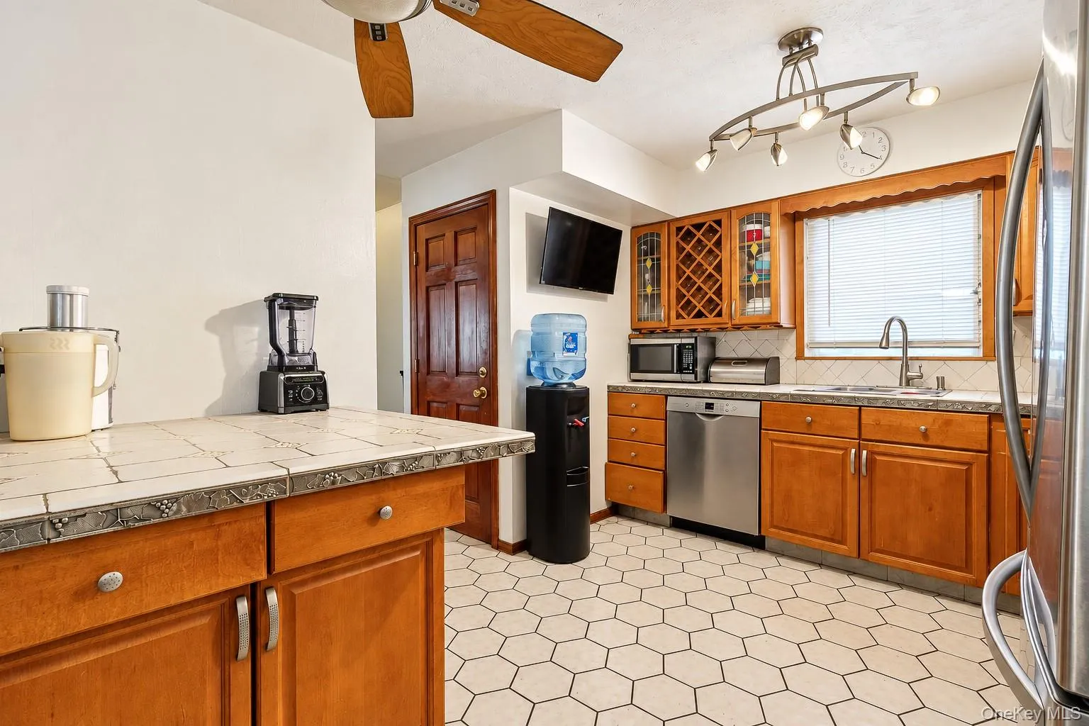 Kitchen featuring tile-topped cabinetry, stainless steel dishwasher, wood-finish cabinetry with integrated wine storage, hexagonal tile flooring, and multi-bulb track lighting Kitchen featuring tile-topped cabinetry, stainless steel dishwasher, wood-finish cabinetry with integrated wine storage, hexagonal tile flooring, and multi-bulb track lighting